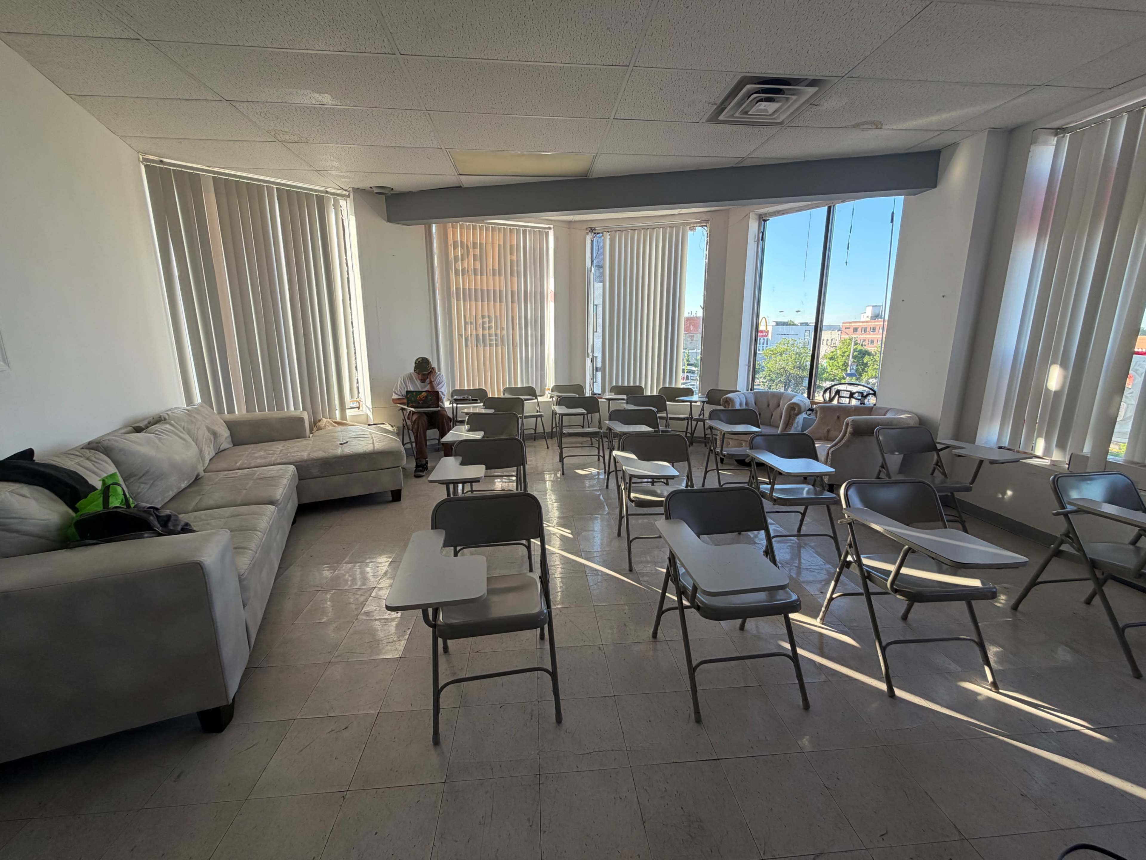The image shows a classroom with rows of empty desks, a sofa in the corner, and a person sitting nearby reading by the window.