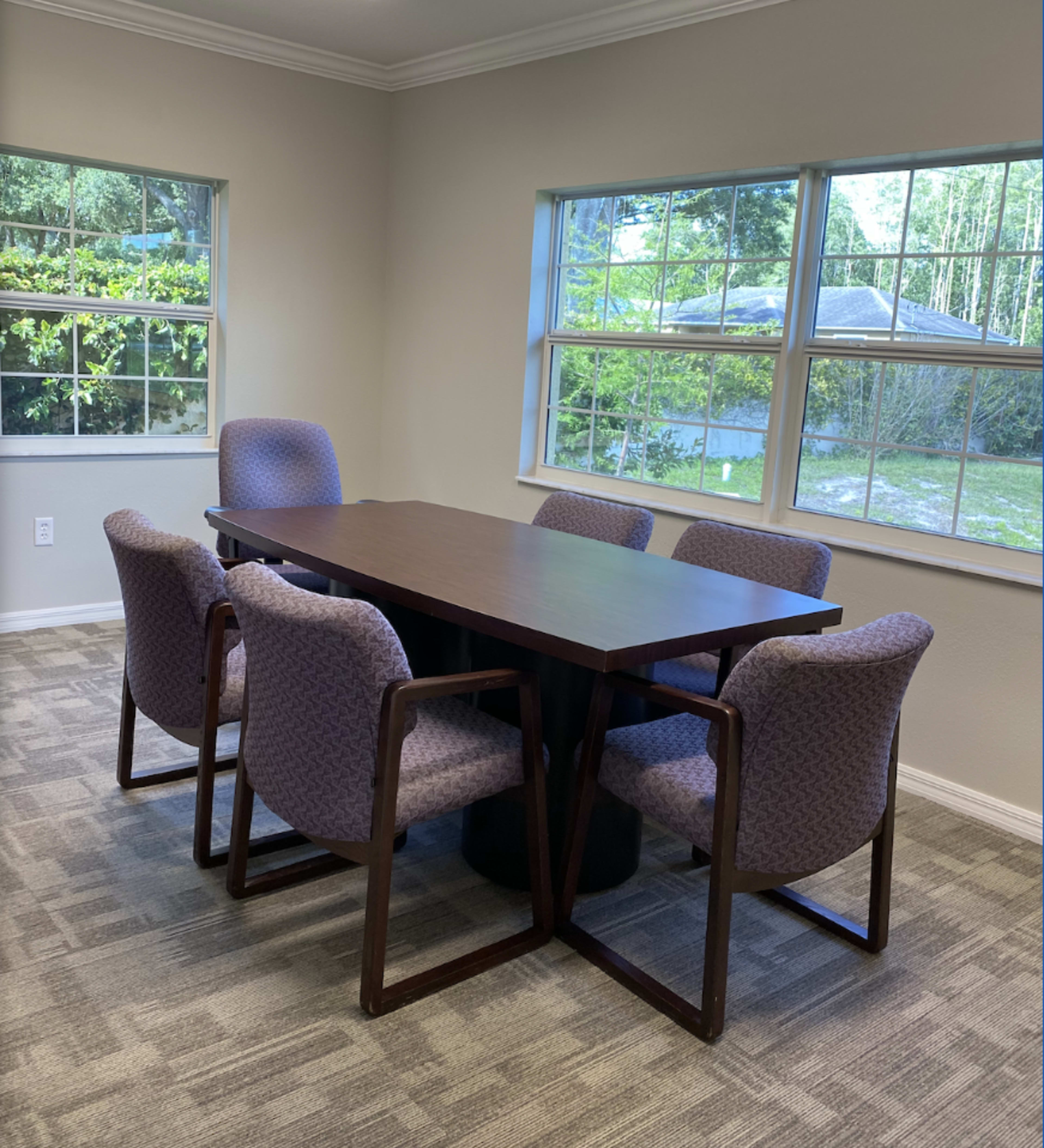 A rectangular wooden table surrounded by six upholstered chairs is positioned in a well-lit room with two windows overlooking a green area.