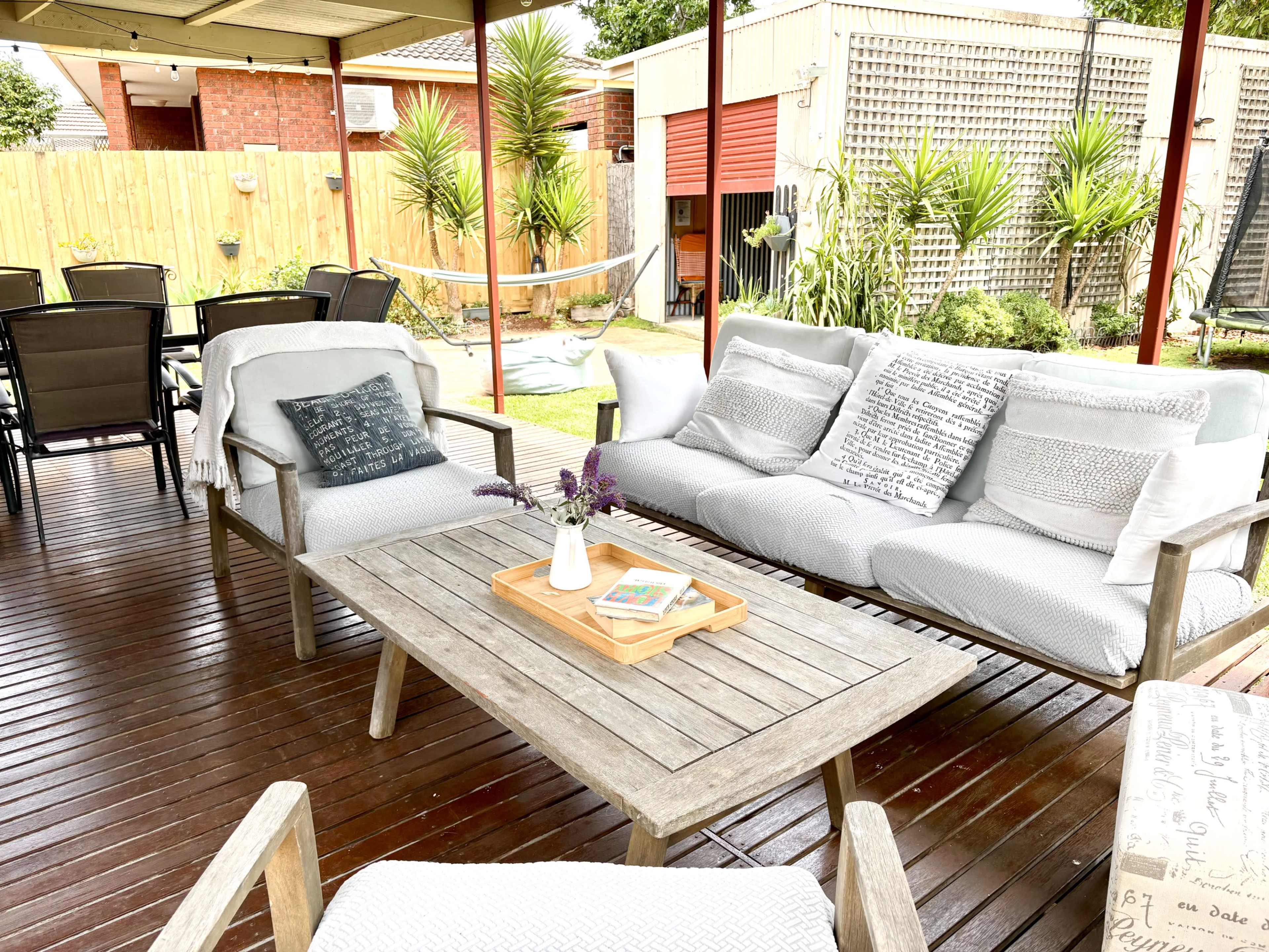 The image shows a cozy outdoor seating area with a wooden table, soft cushions, and greenery in the background, including a hammock and patio furniture.