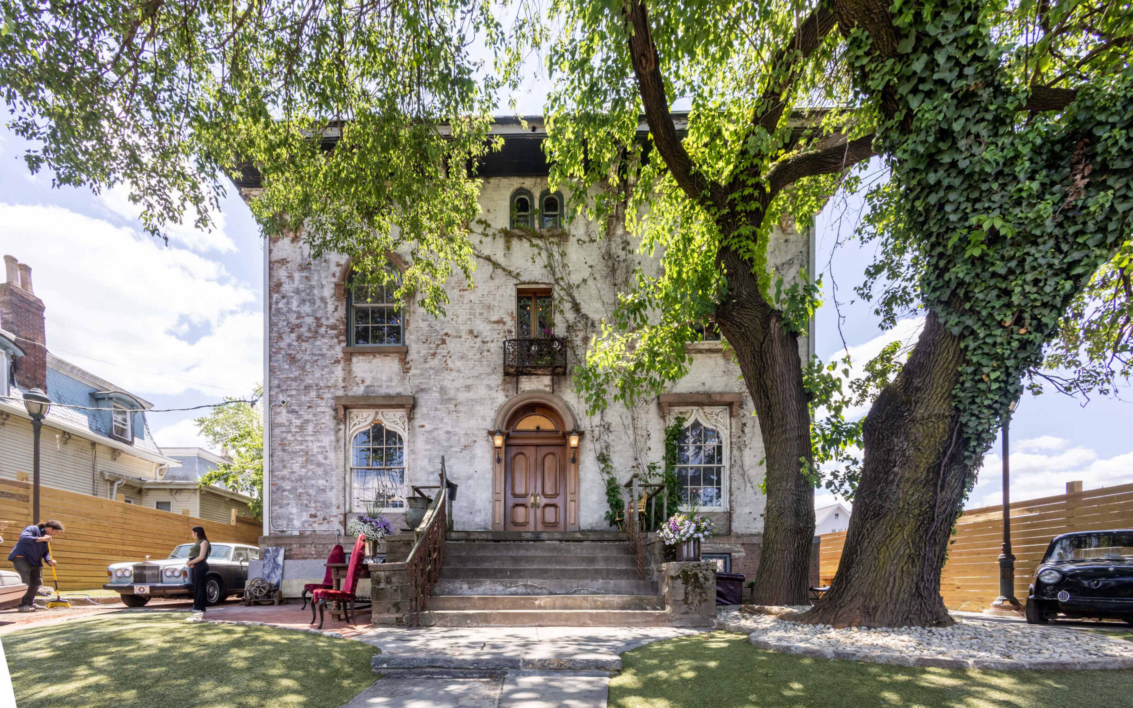 A historic two-story brick house with a large front staircase, surrounded by trees and greenery.