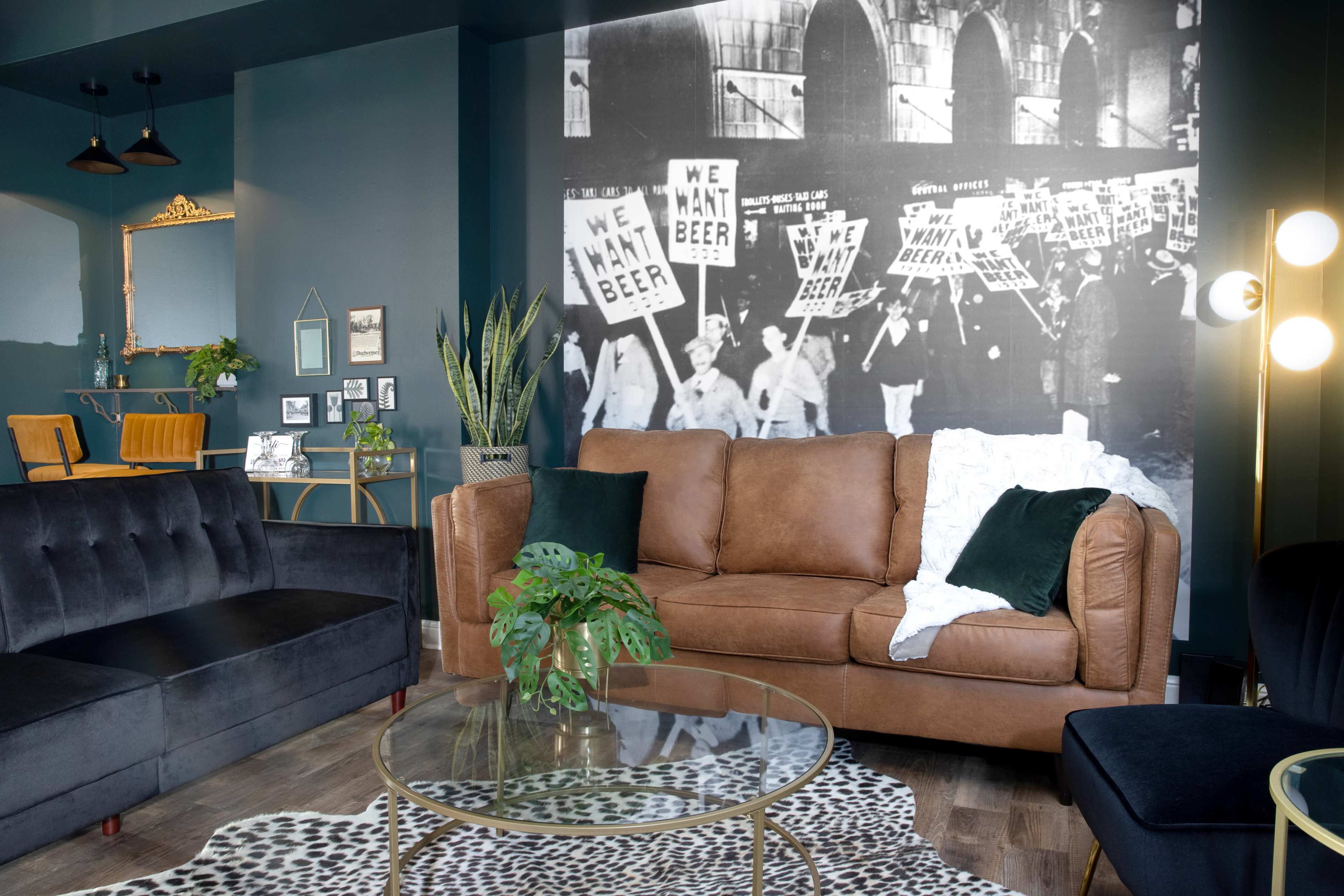 The image features a living room with a brown leather sofa, a glass coffee table on a leopard print rug, and a large black-and-white mural depicting a historical protest for beer.