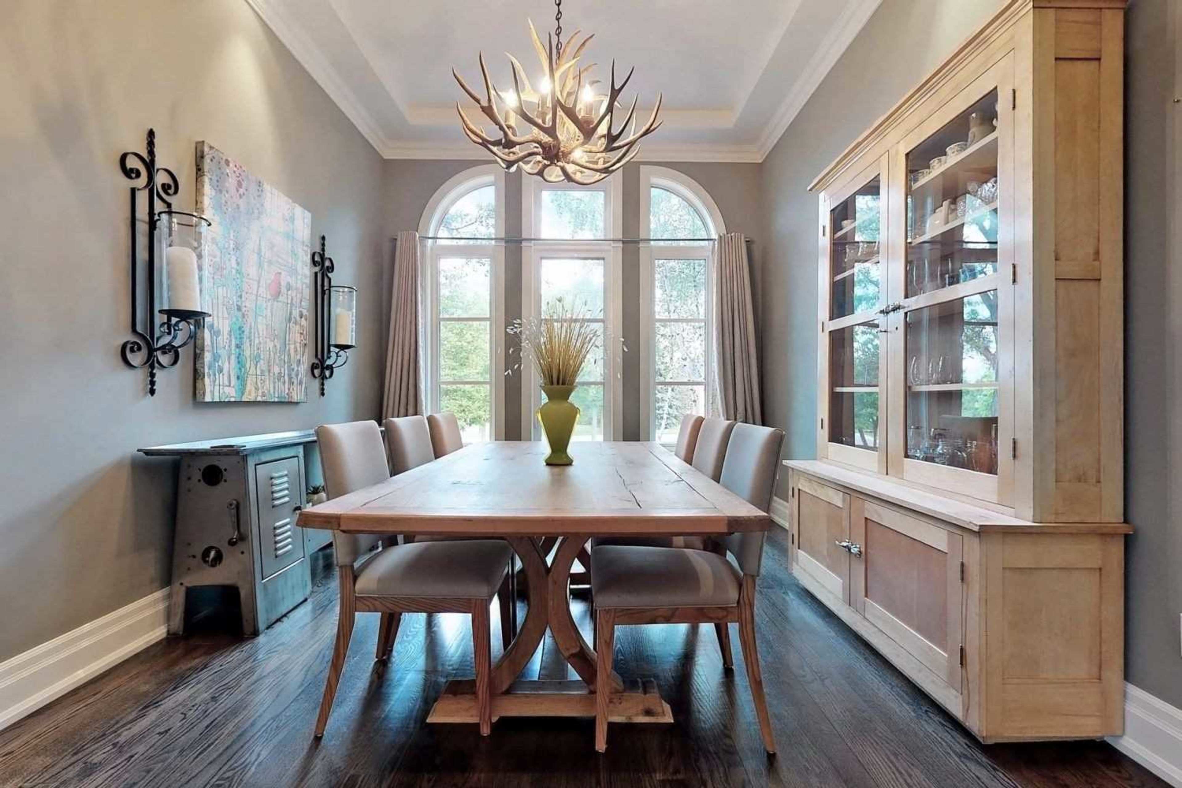 A spacious dining room features a wooden table surrounded by gray upholstered chairs, an antler chandelier above, and large windows allowing natural light to enter.
