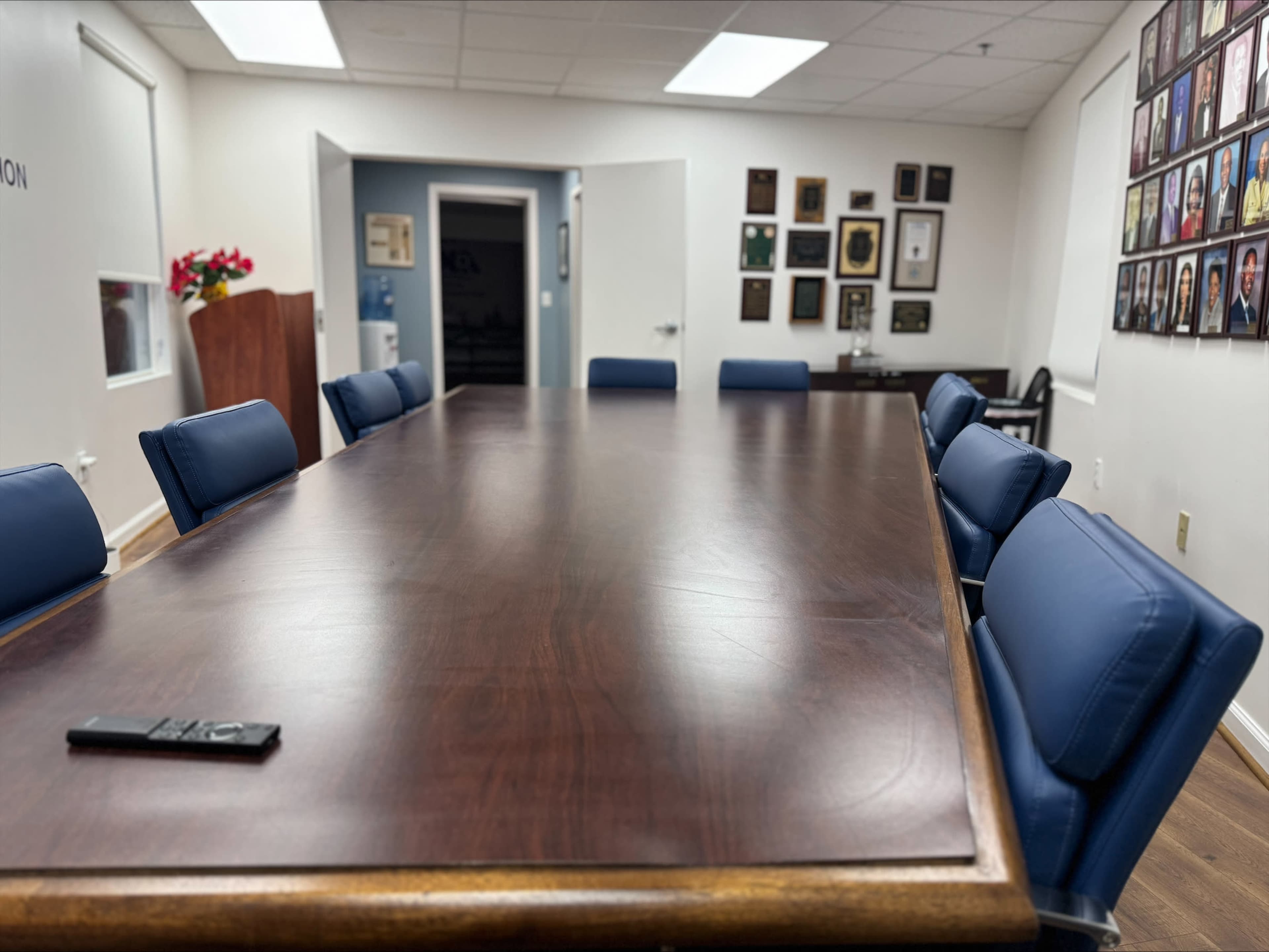 A large, rectangular wooden conference table is set in a well-lit room with blue chairs around it, and walls adorned with framed photographs and certificates.