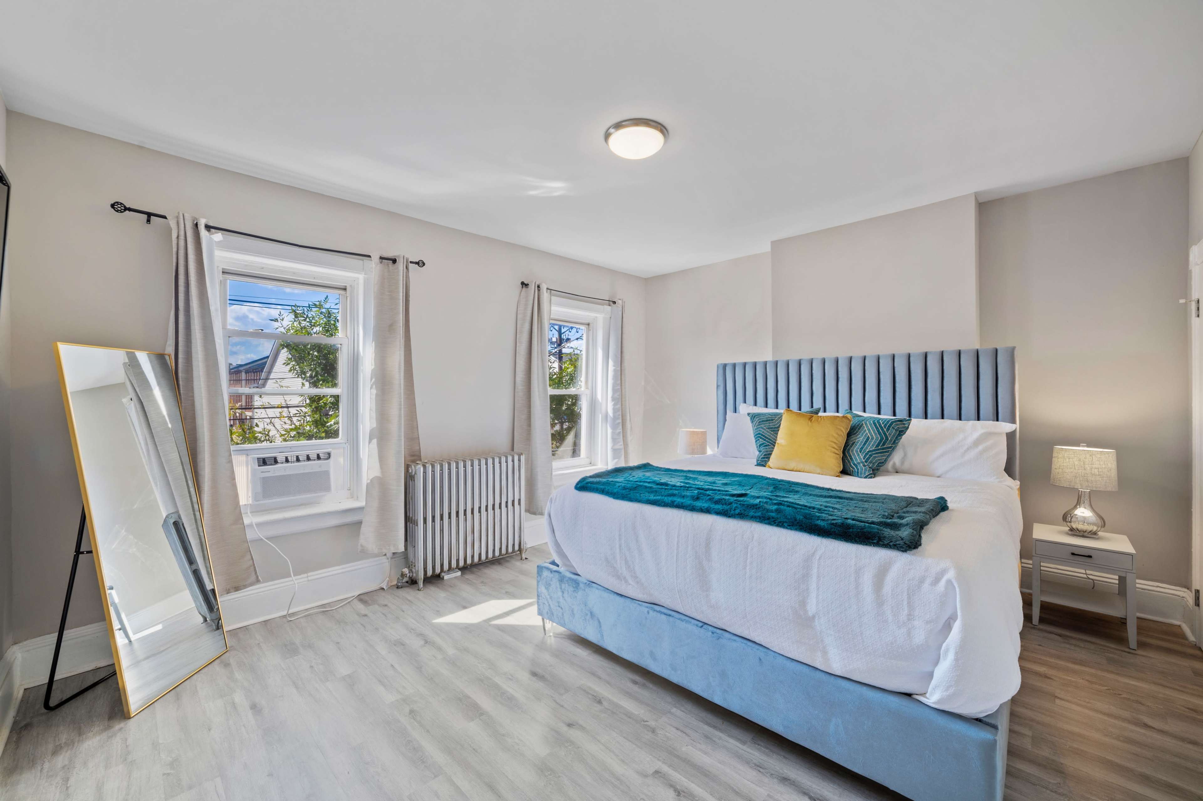 A well-lit bedroom featuring a bed with a blue headboard, a mirror, a nightstand with a lamp, and two windows.