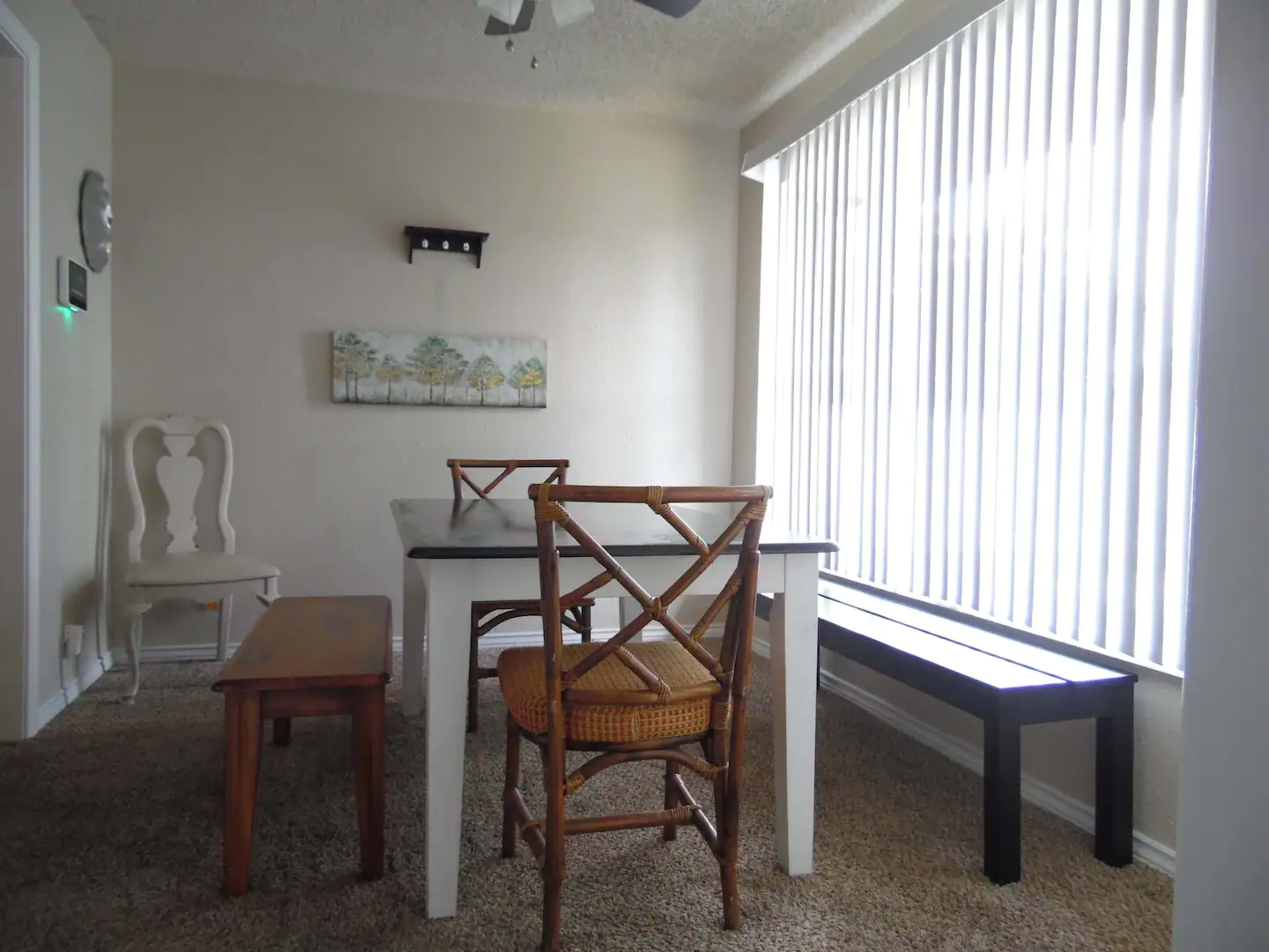 A dining area features a white table with two benches and a chair, set against a wall of vertical blinds and artwork.