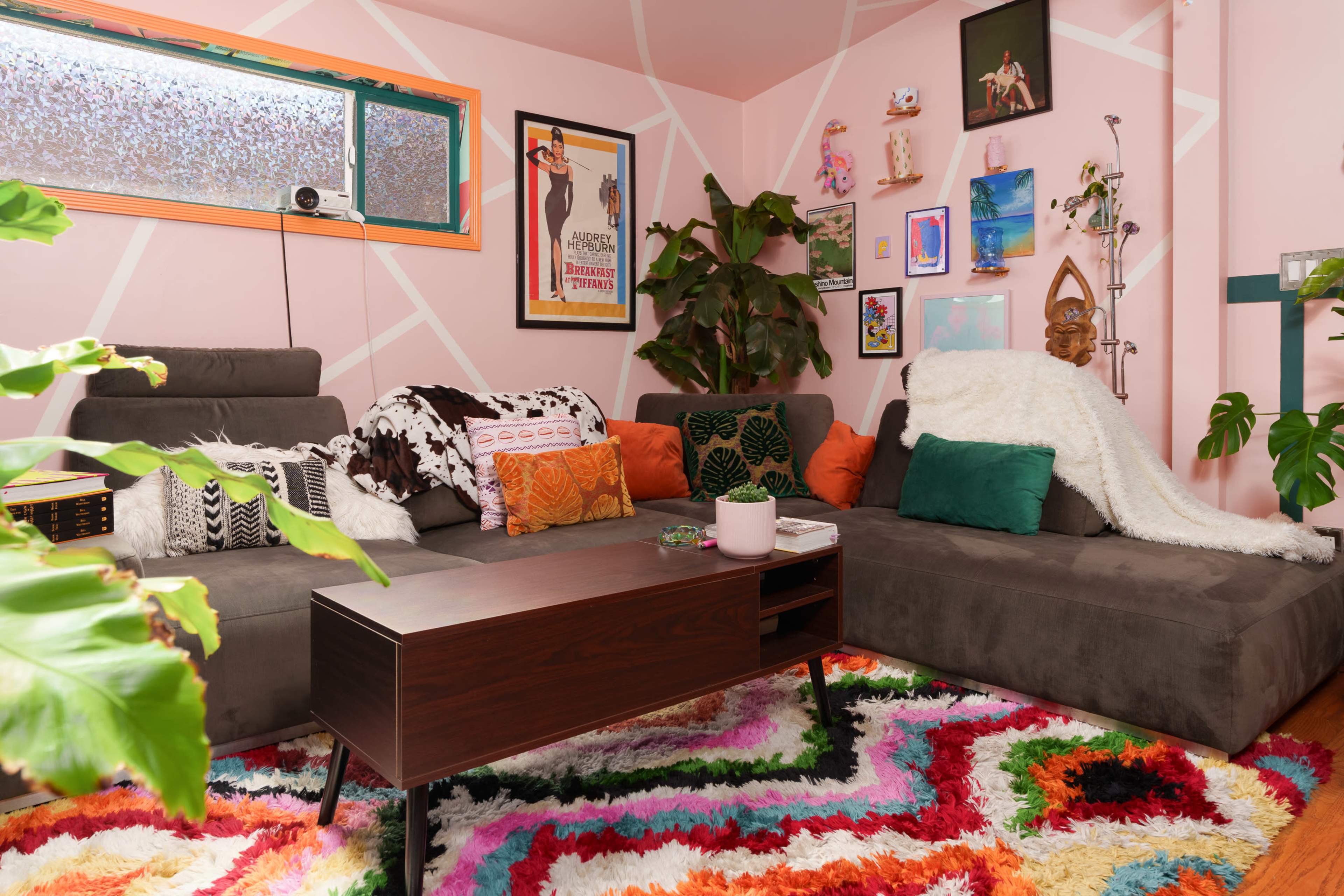 The image shows a vibrant living room with a gray sectional sofa adorned with colorful pillows, a wooden coffee table, and artwork on the walls, set against a pink background with geometric patterns.