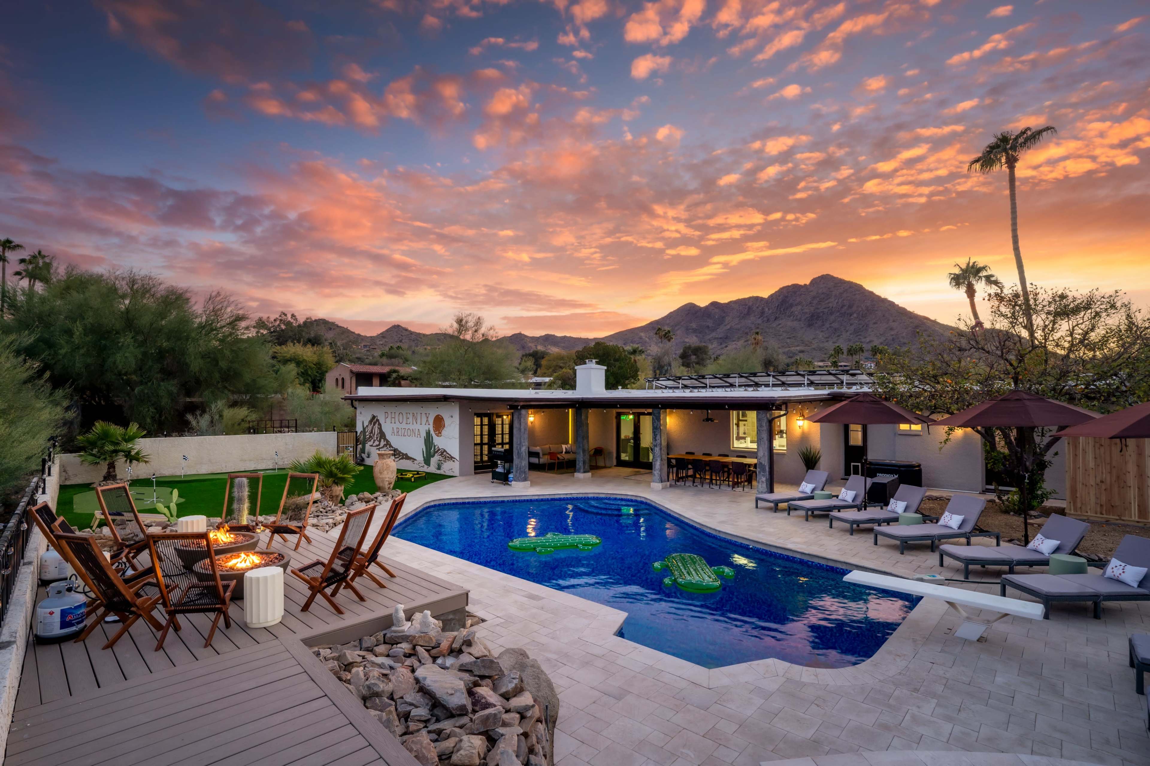 The scene features a tranquil outdoor area with a swimming pool, surrounded by lounge chairs and an outdoor dining setup, set against a colorful sunset sky and mountains in the background.