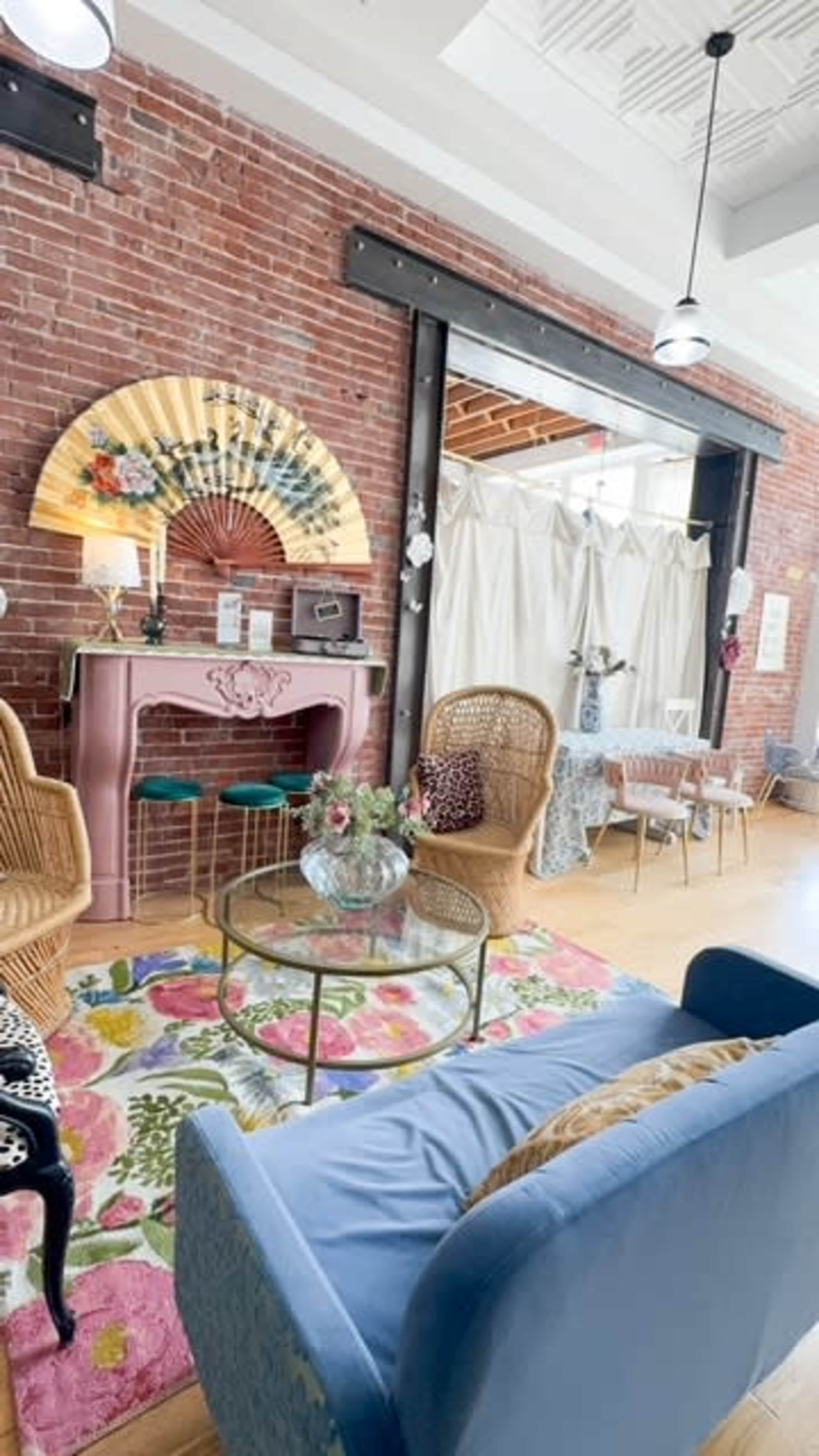 The image depicts a stylish interior of a café or boutique featuring a brick wall, a pink vintage console, wicker seating, and a colorful floral rug.