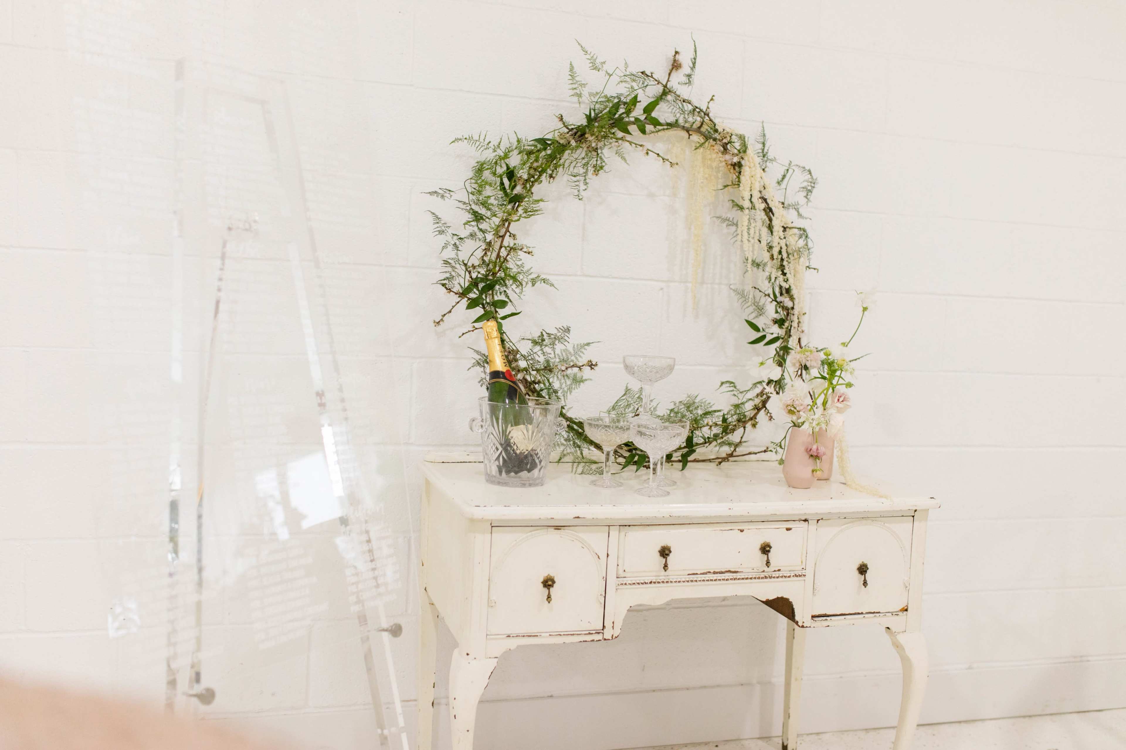 A vintage white table is adorned with a circular floral wreath, a champagne bottle in an ice bucket, a glass bowl, and a small vase of flowers.