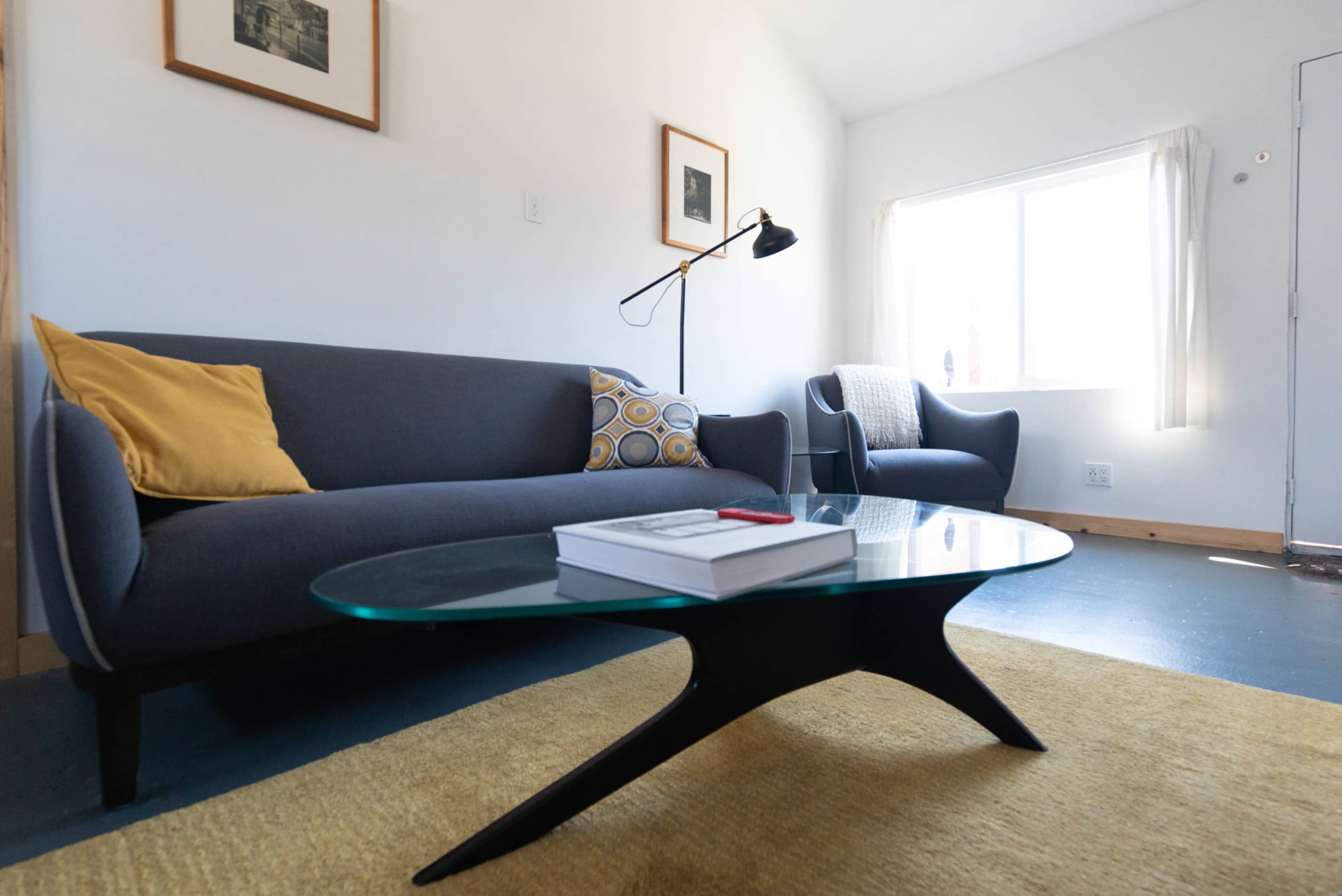 The image shows a modern living room featuring a gray sofa with yellow and patterned pillows, a glass coffee table, and two armchairs near a window.