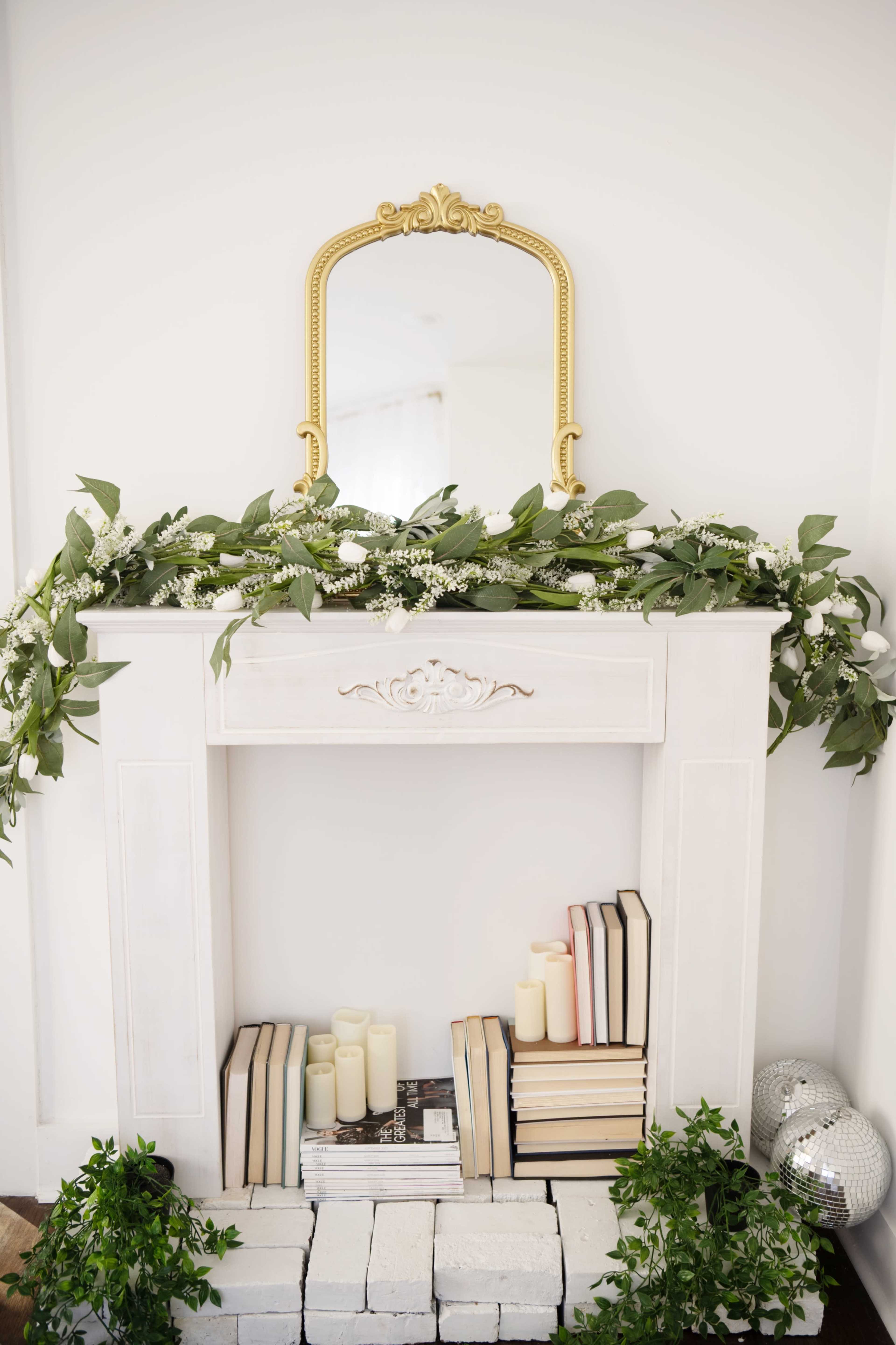 A white mantelpiece adorned with greenery and a gold-framed mirror sits against a light wall, featuring candles and stacked books on the ledge below.