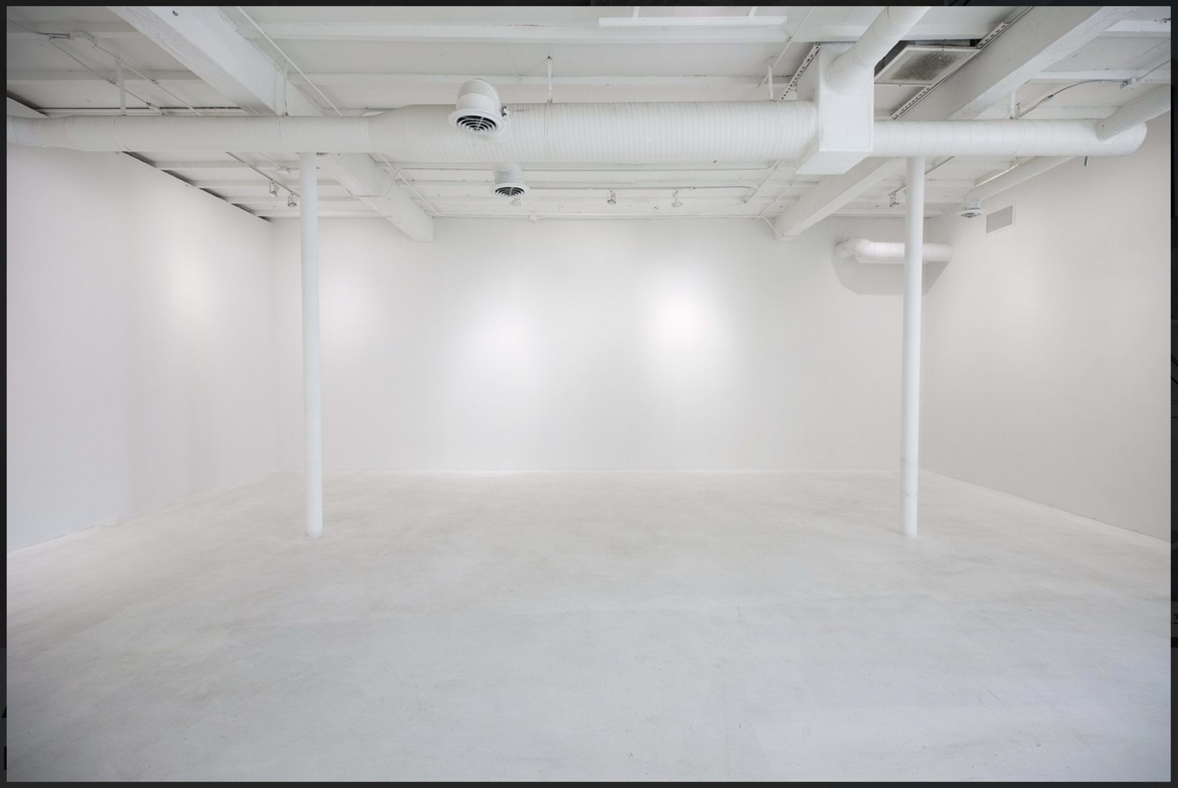 The image shows a spacious, empty room with white walls and a white floor, featuring several columns and overhead ventilation ducts.