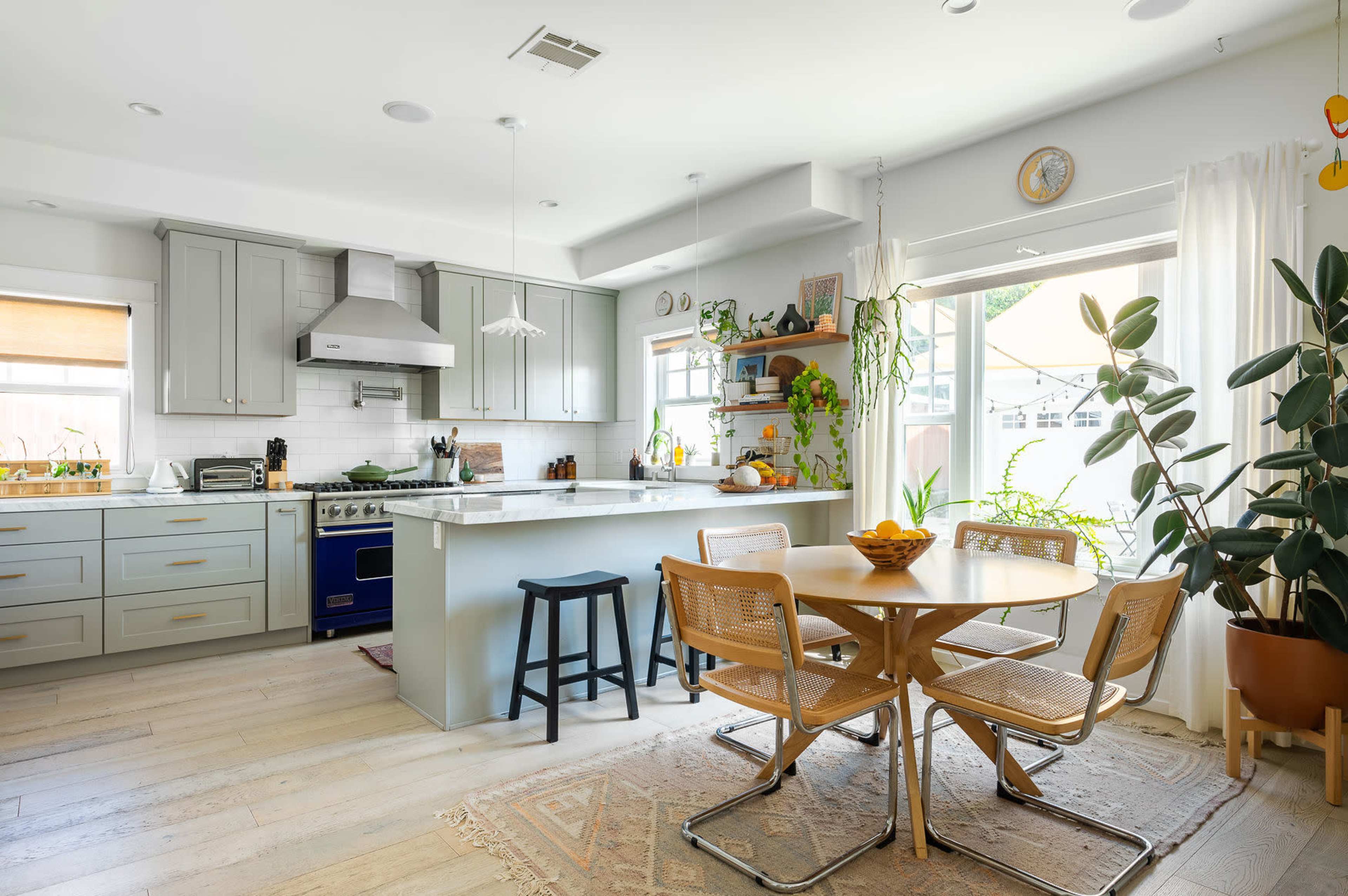 A modern kitchen with gray cabinets, a central island, a round dining table with four chairs, and various houseplants.