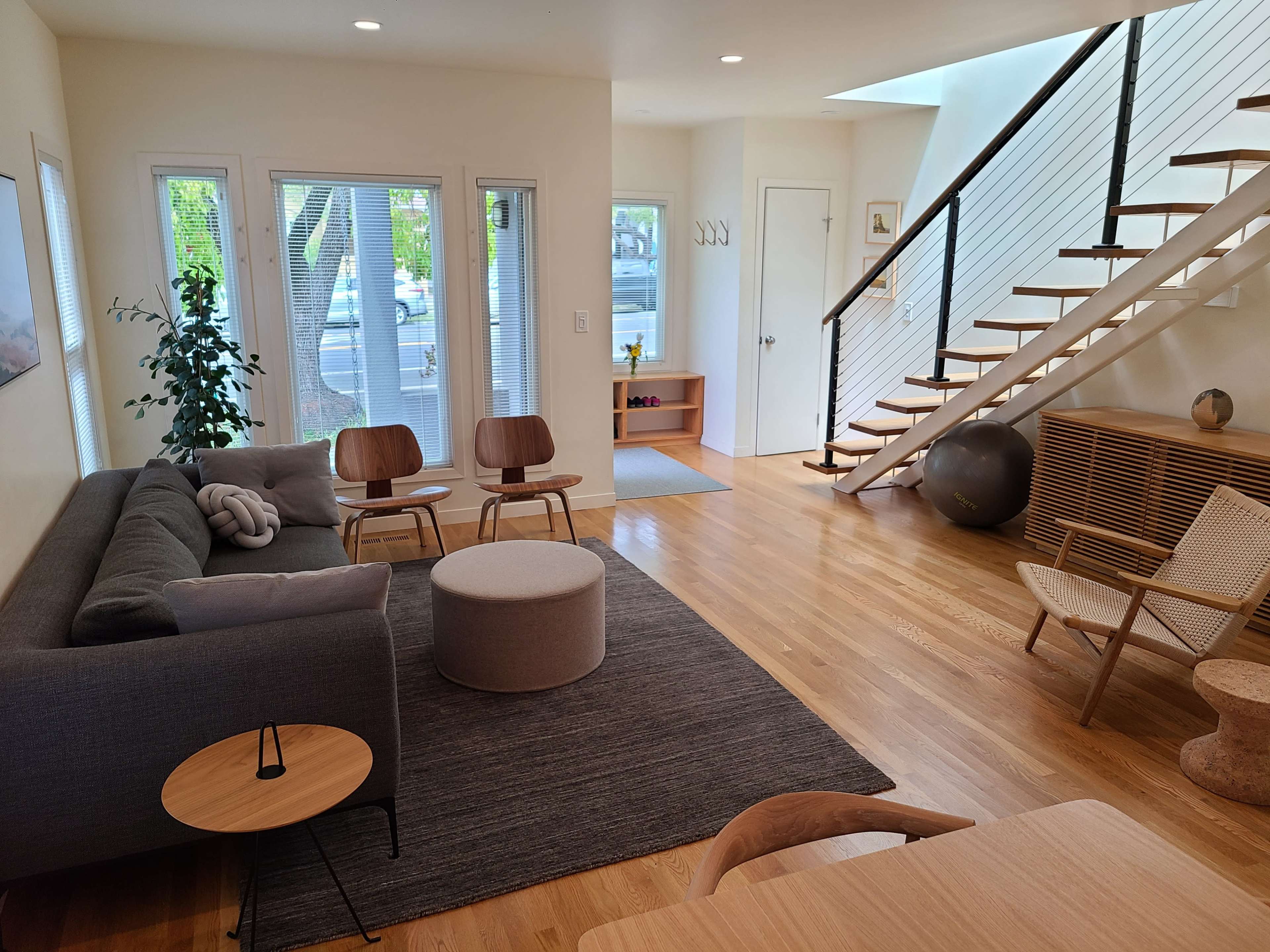 A modern living room featuring a gray sofa, two wooden chairs, a round ottoman, and a staircase with a wooden railing.