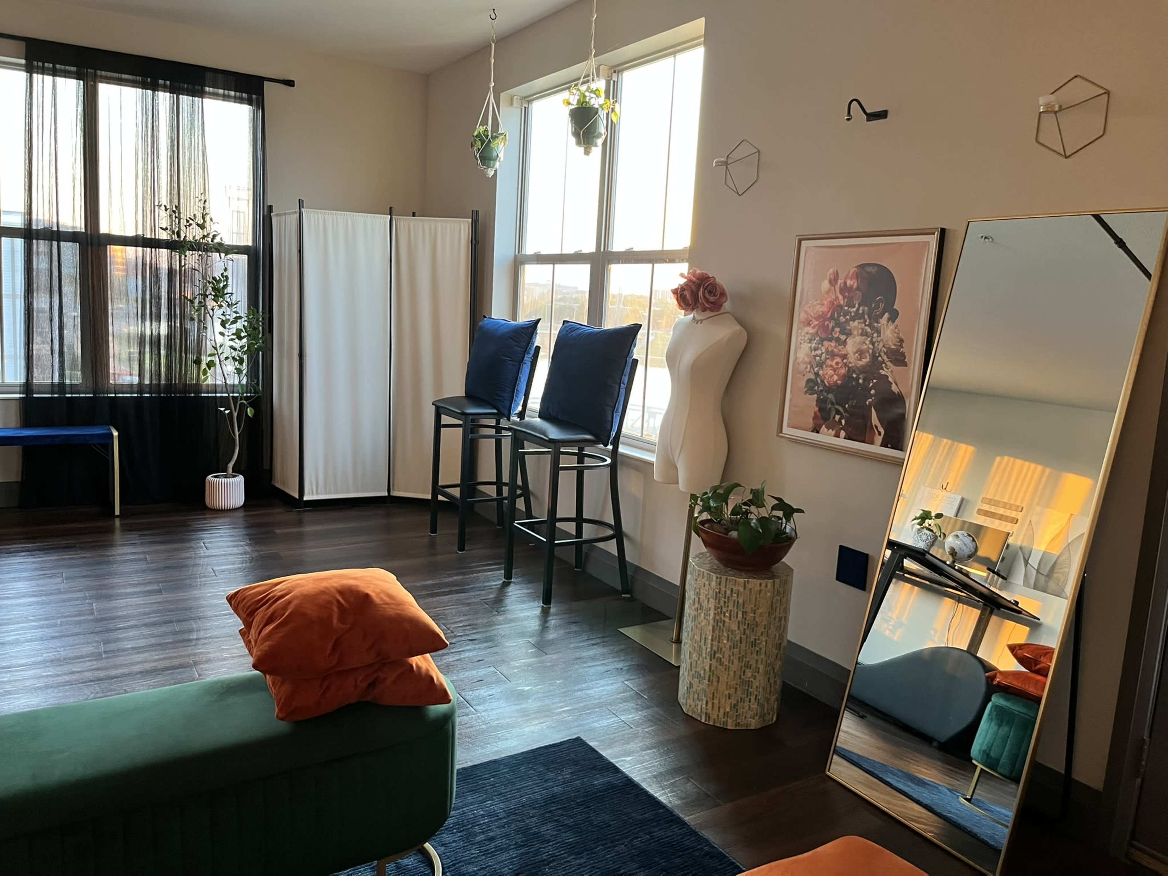 The image shows a modern interior space featuring a green sofa, a large mirror, a clothing mannequin, potted plants, and decorative artwork, with large windows allowing natural light to enter.
