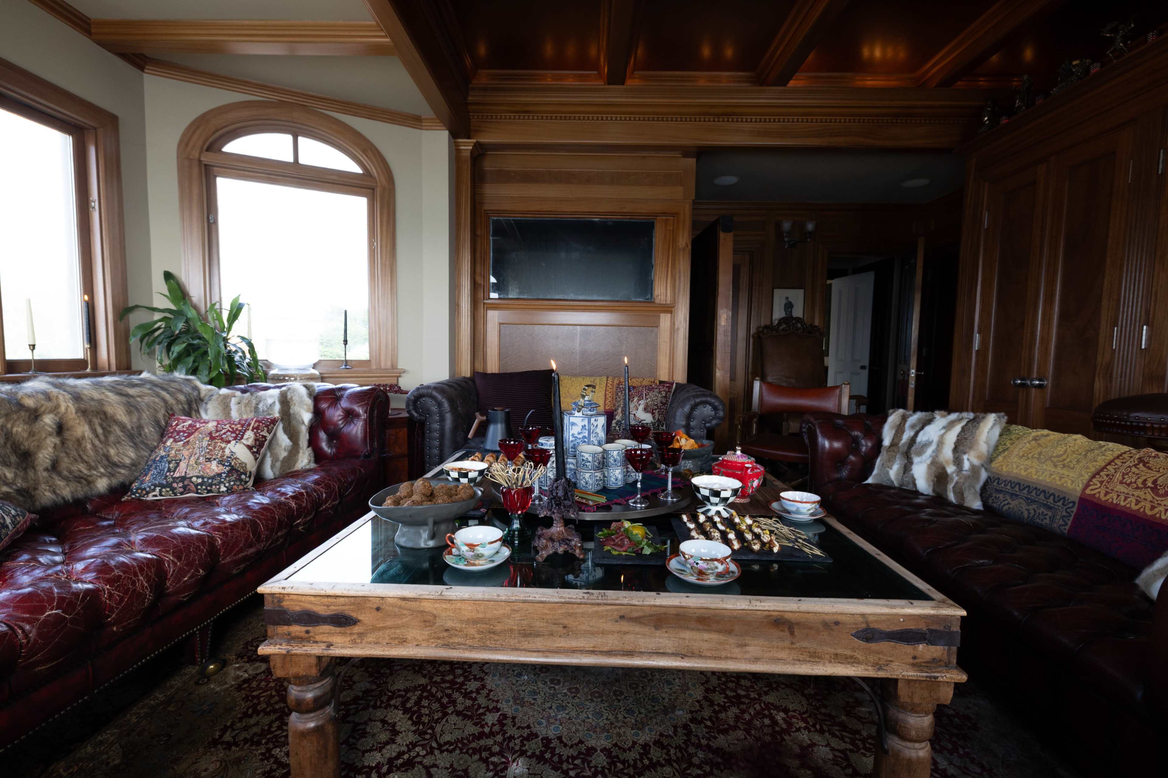 The image shows a richly furnished living room with dark leather sofas, a wooden coffee table adorned with snacks and decorative items, and large windows allowing natural light to enter.