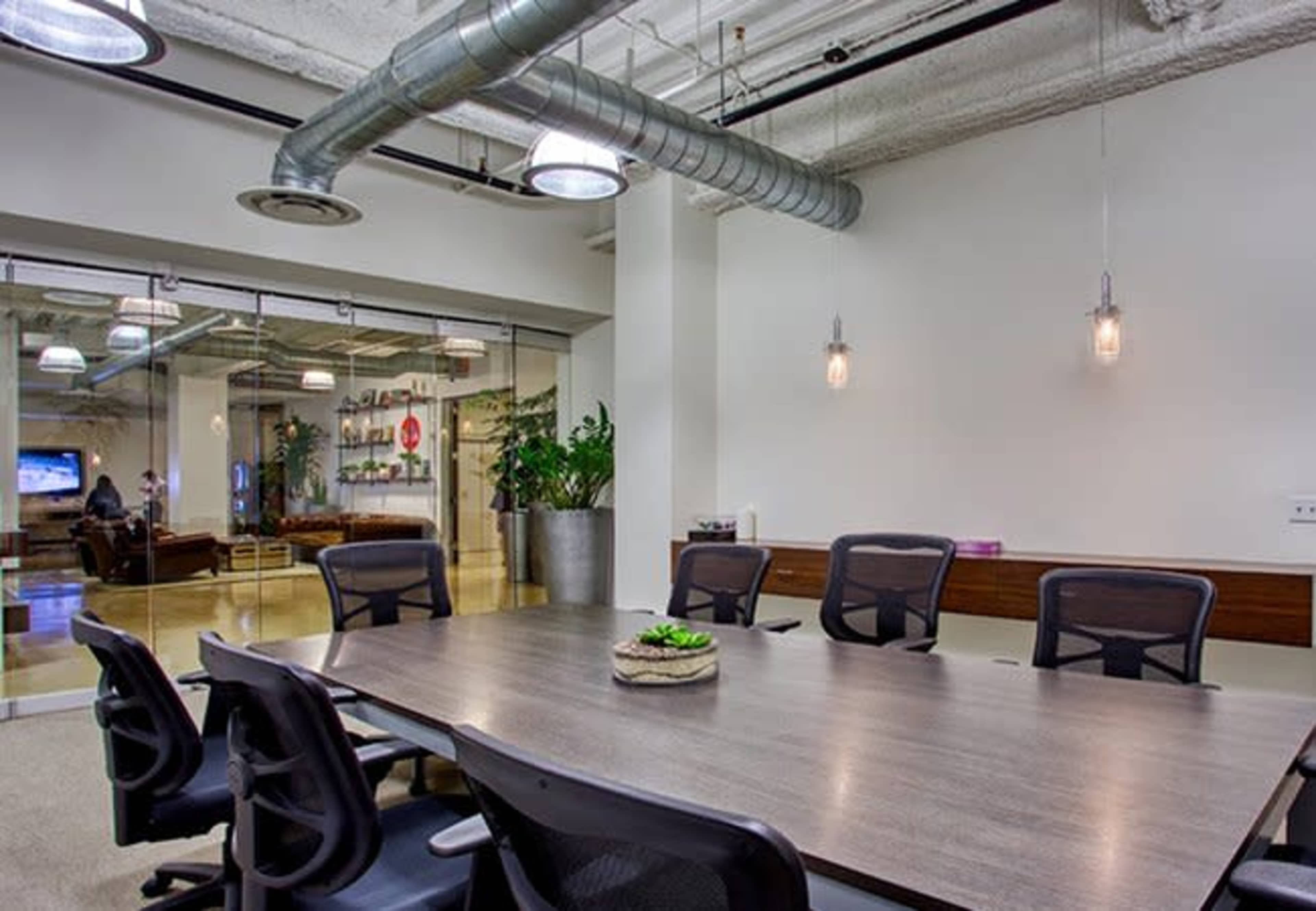 A modern conference room features a large wooden table surrounded by black chairs, with glass walls revealing a seating area beyond.