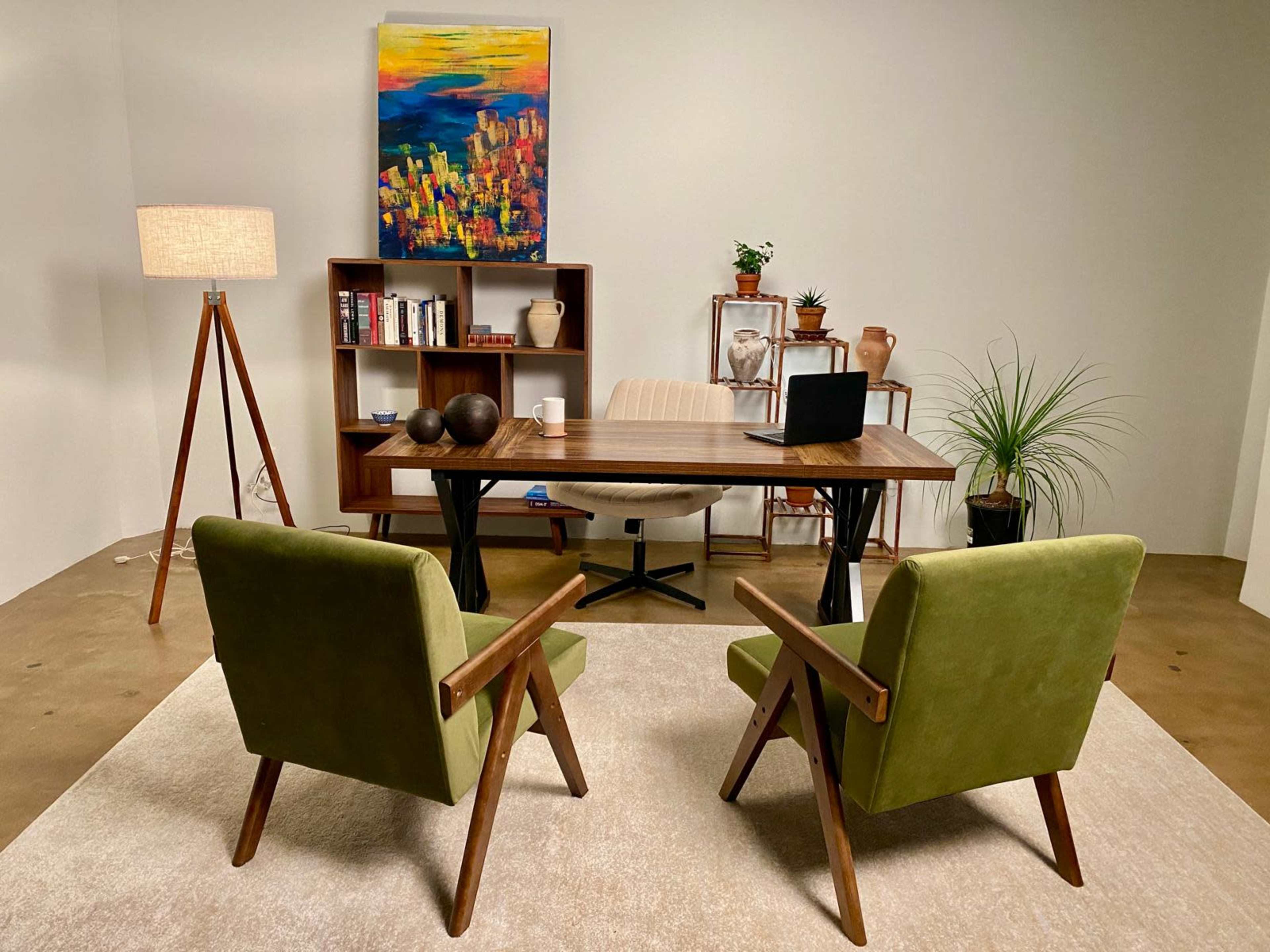 A modern office interior features a wooden desk with a laptop, two green chairs, a floor lamp, and shelves displaying books and plants.