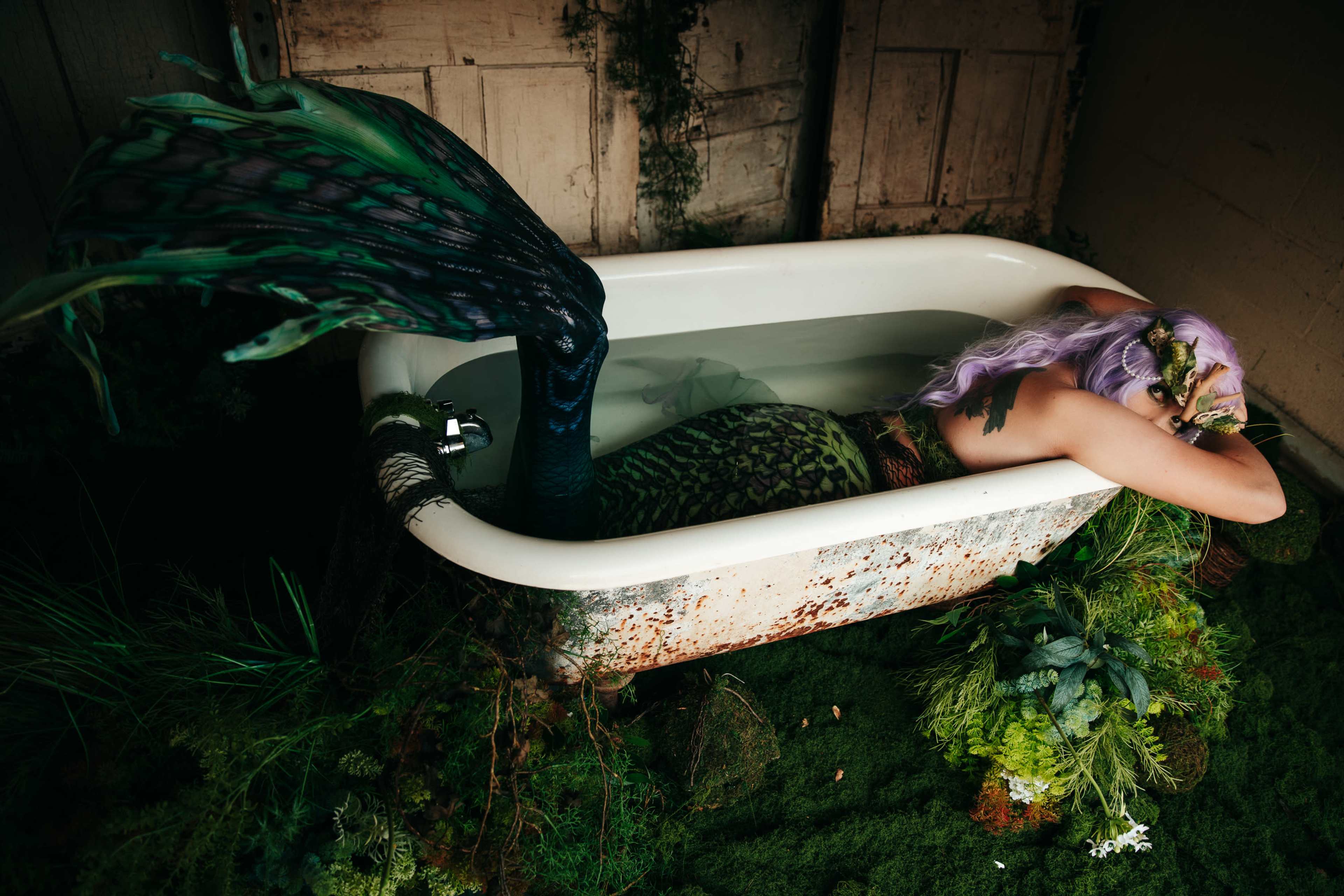 A person with a mermaid tail rests in a vintage bathtub surrounded by lush greenery and moss.