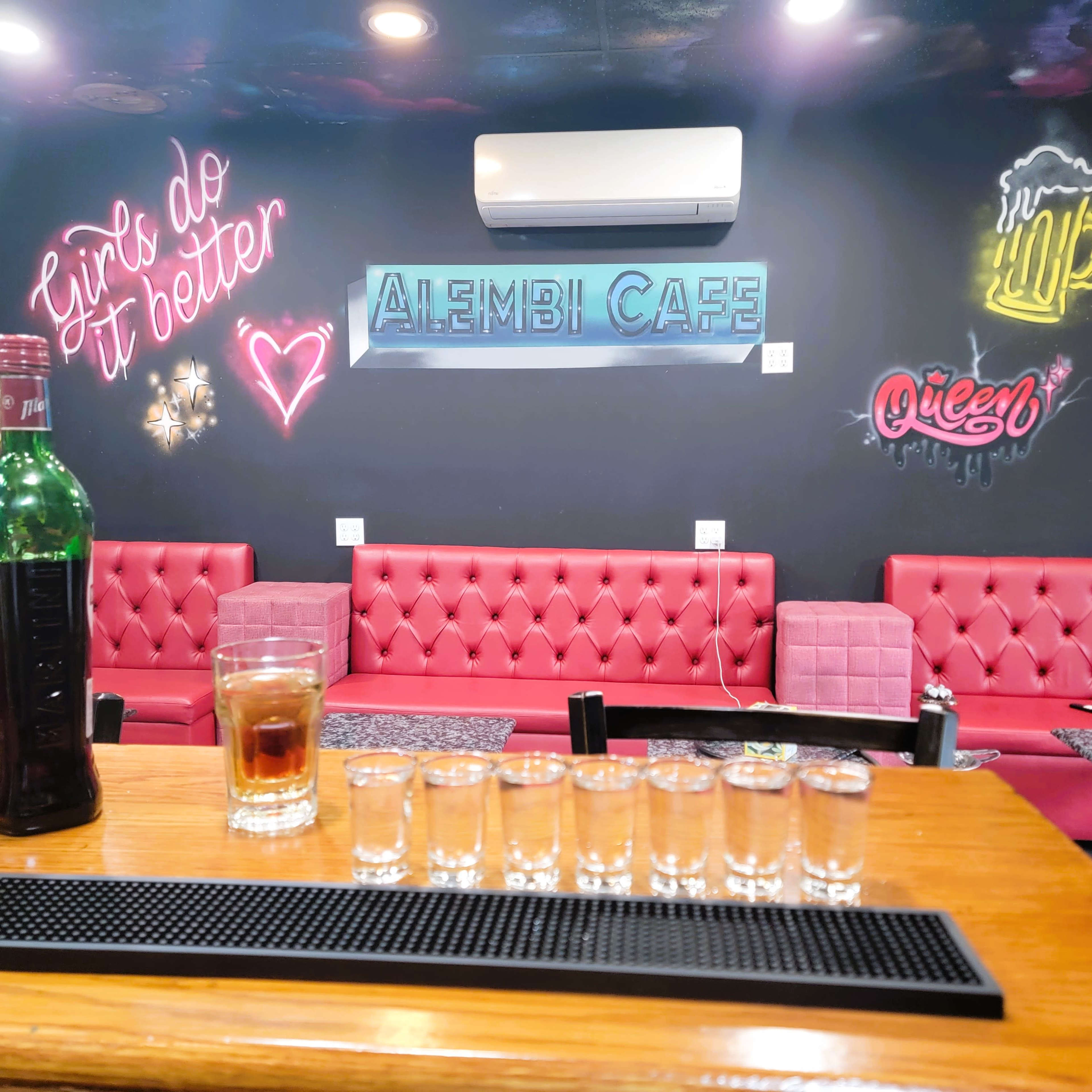 The image shows a bar with a wooden countertop featuring several shot glasses lined up in front of a glass of a dark beverage and a bottle, with a colorful mural on the wall behind it.
