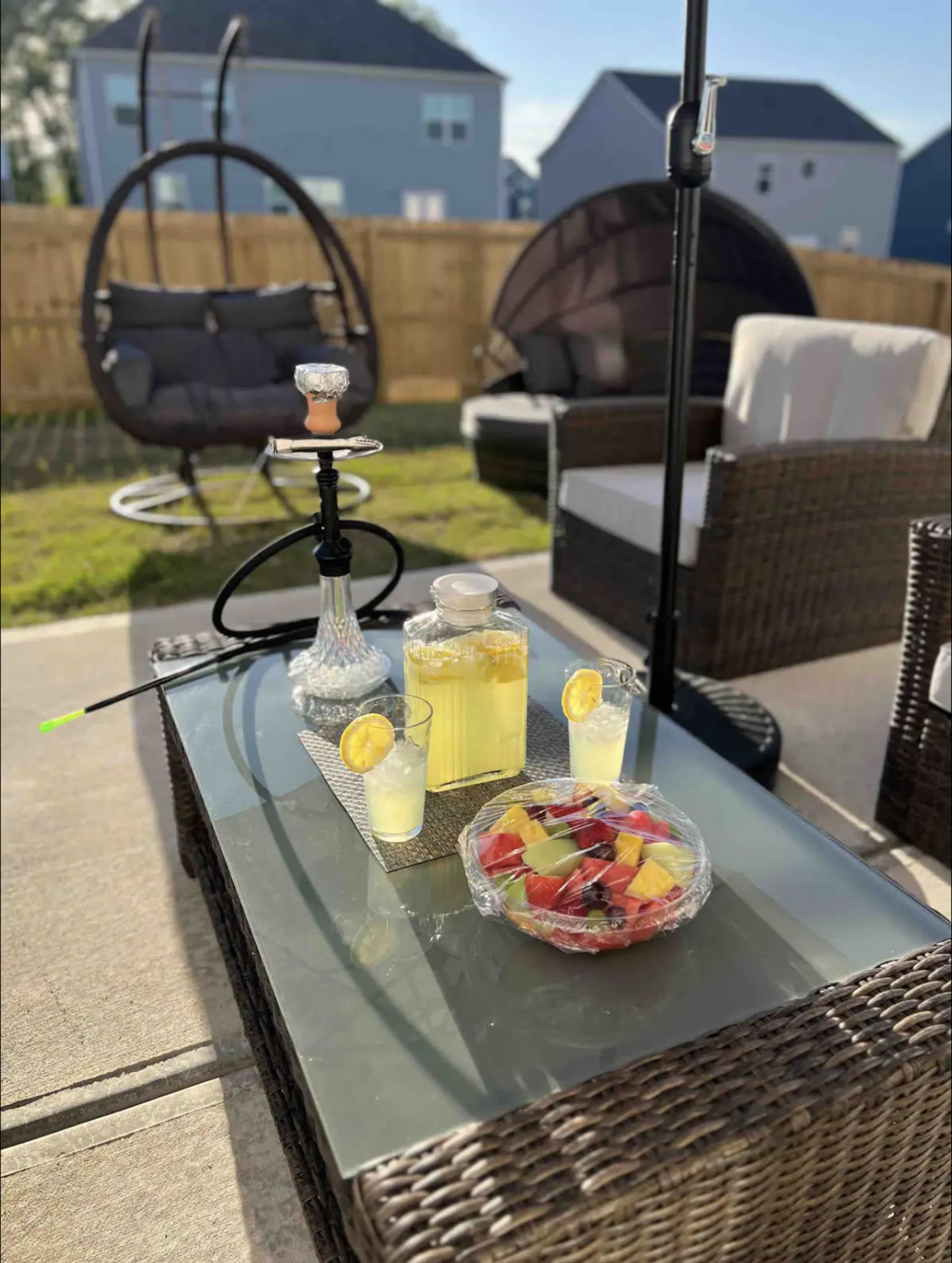 A glass table holds a pitcher of lemonade, two glasses, a hookah, and a bowl of fruit, with outdoor furniture and a wooden fence in the background.