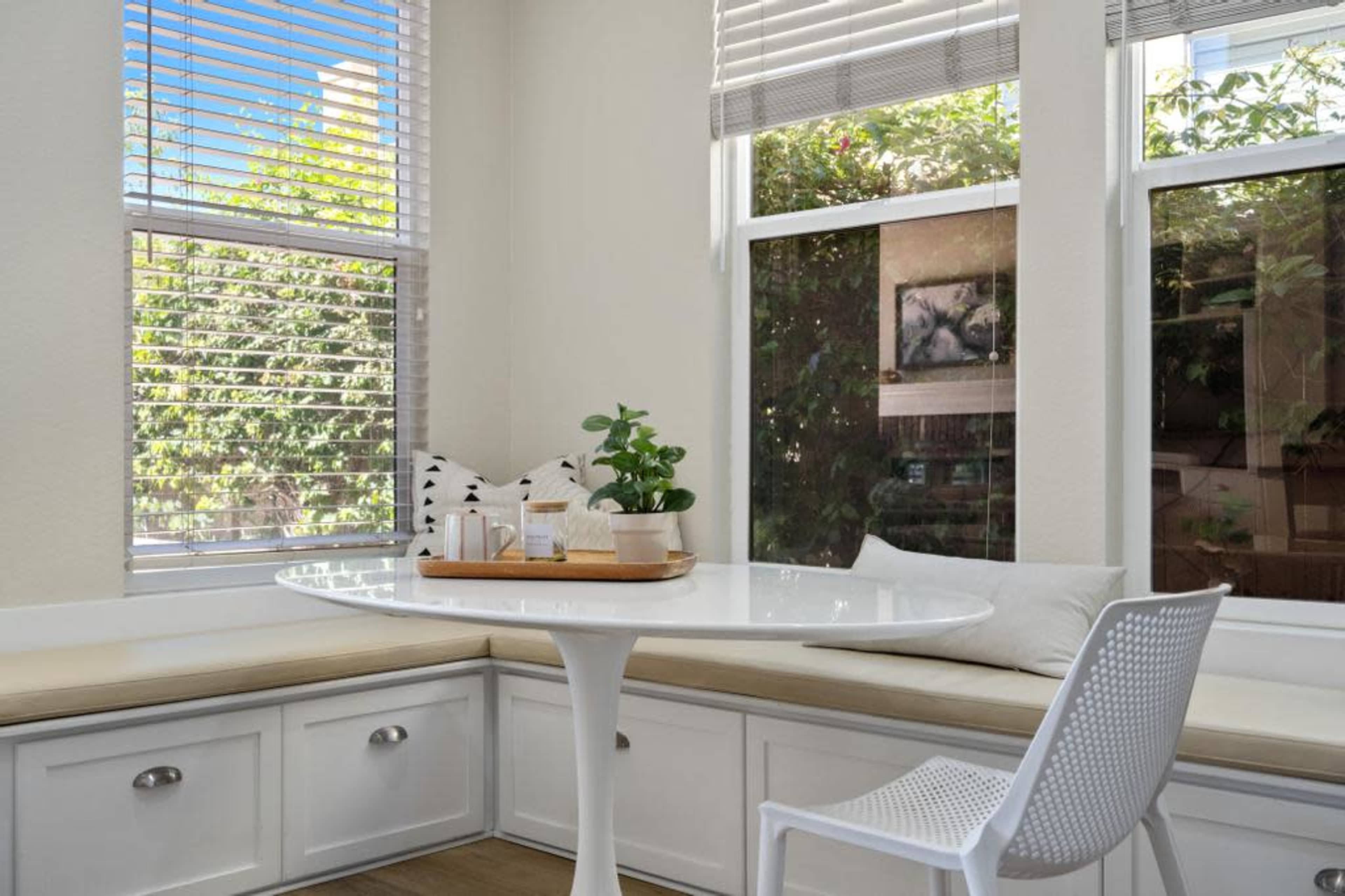 A round white table is placed in a bright corner next to large windows with greenery visible outside.