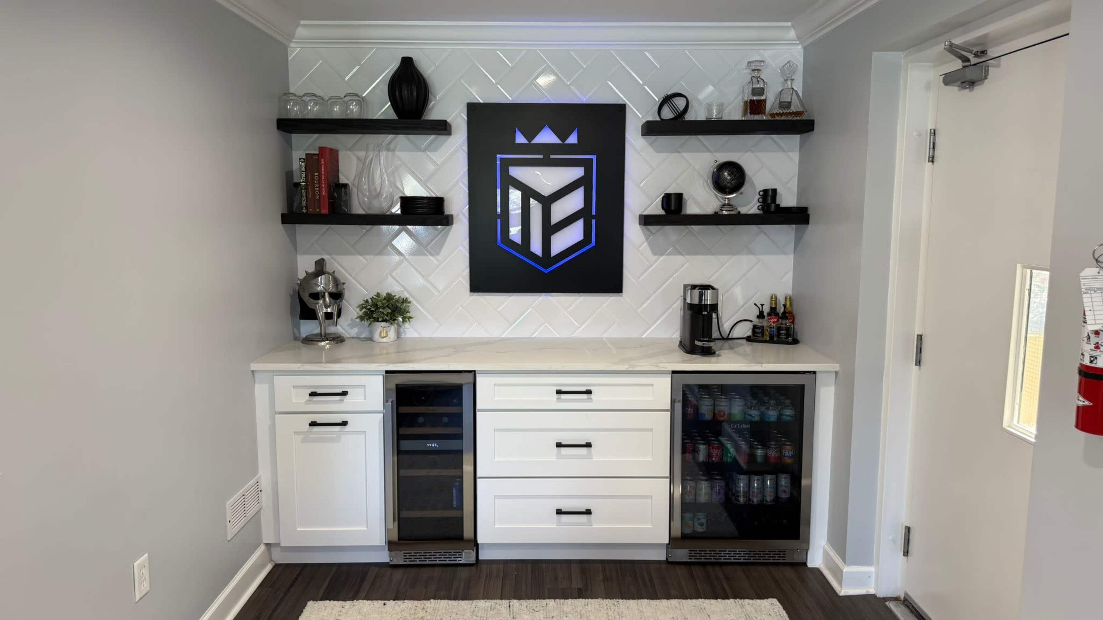 The image shows a modern kitchenette with white cabinetry, a marble countertop, a wall with decorative shelves, and a black and blue emblem displayed prominently.