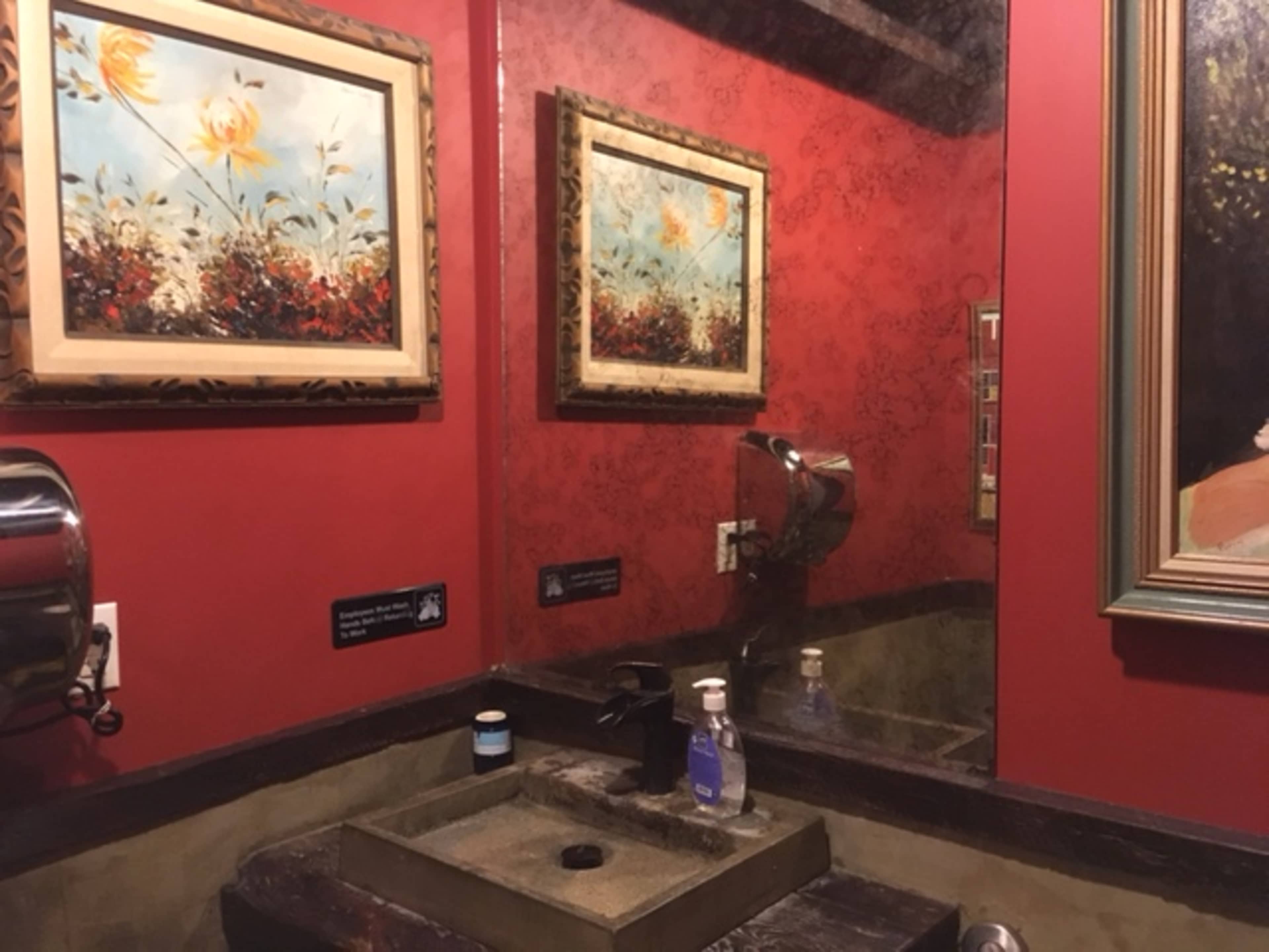 A small restroom with a sink, wall-mounted hand dryer, and two framed artworks hanging on red-painted walls.