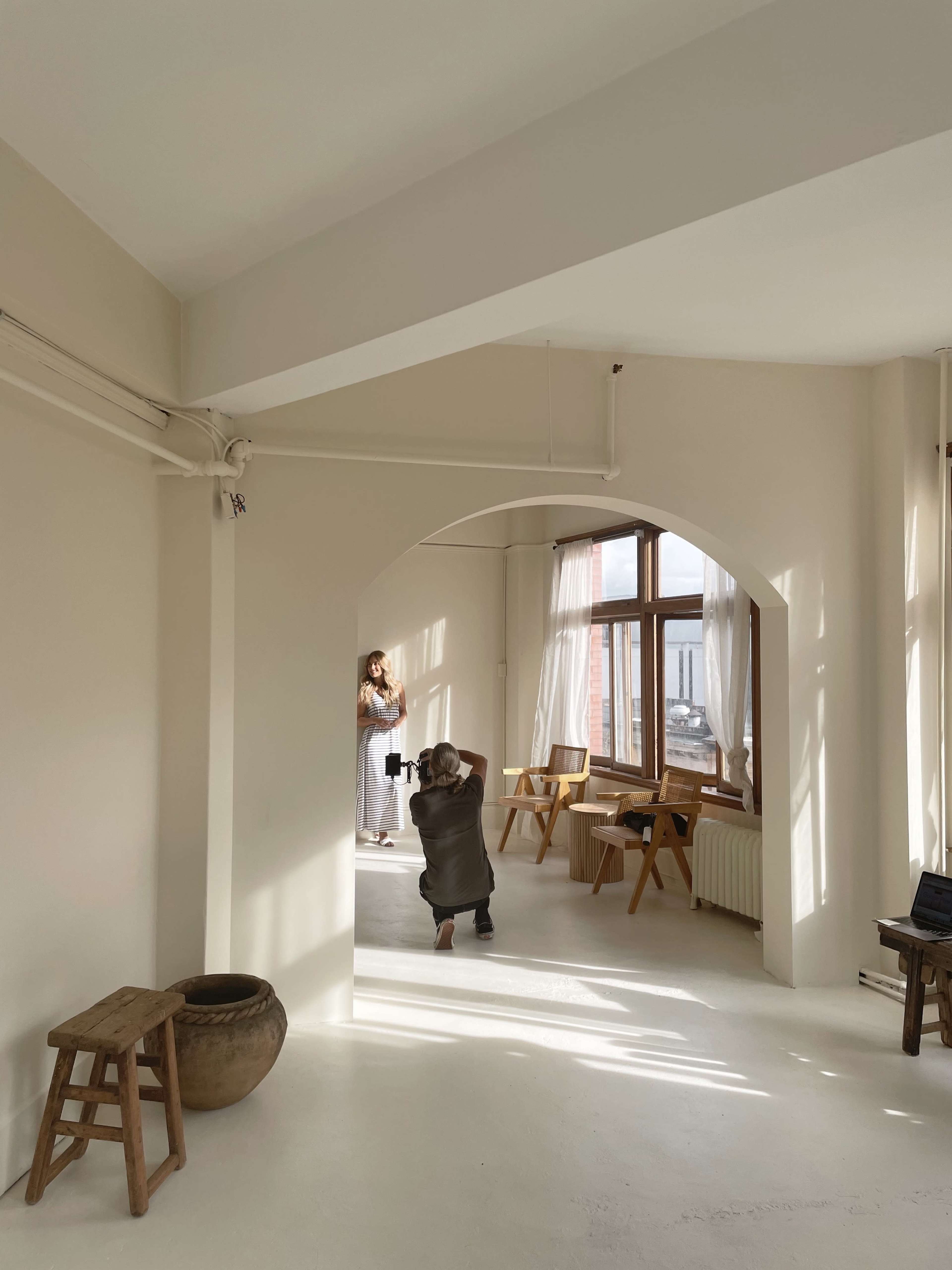 A photographer captures an image of a model in a bright, airy room with large windows and minimal furnishings.
