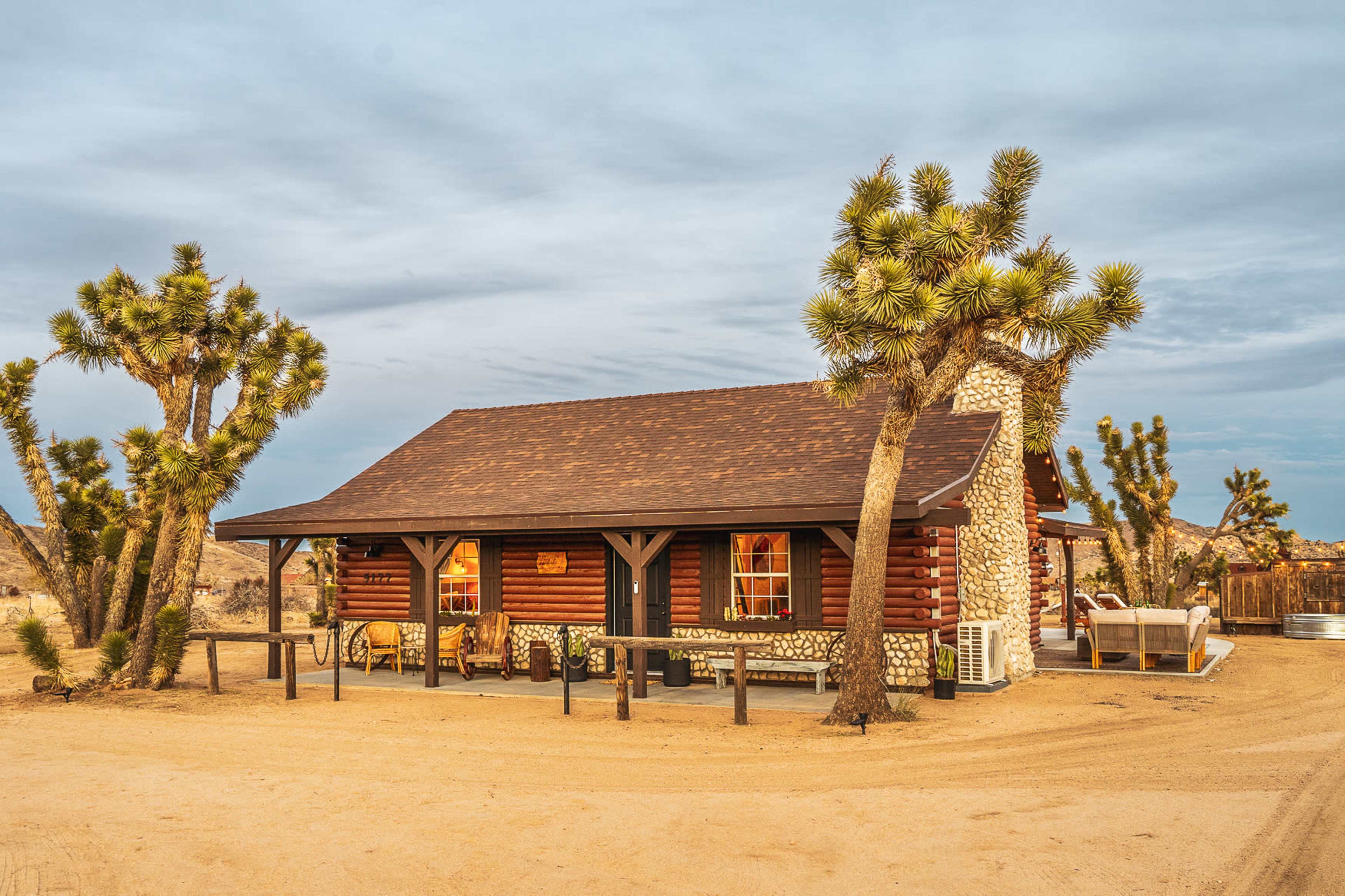 A log cabin with a stone chimney is surrounded by Joshua trees and sits on a sandy landscape under a cloudy sky.