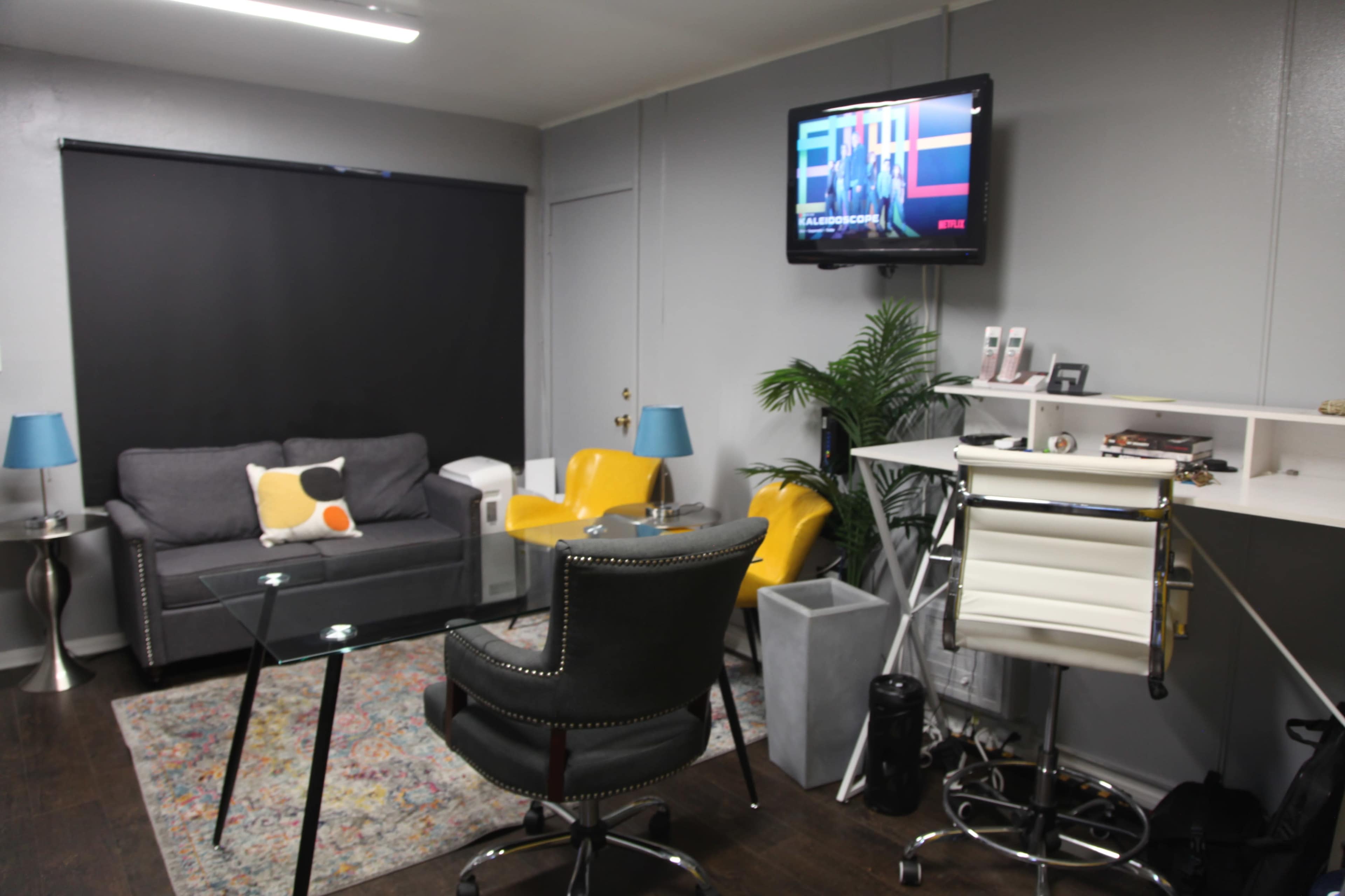 The room features a gray sofa, two yellow chairs, a glass table, a desk with a rolling chair, and a television mounted on the wall.