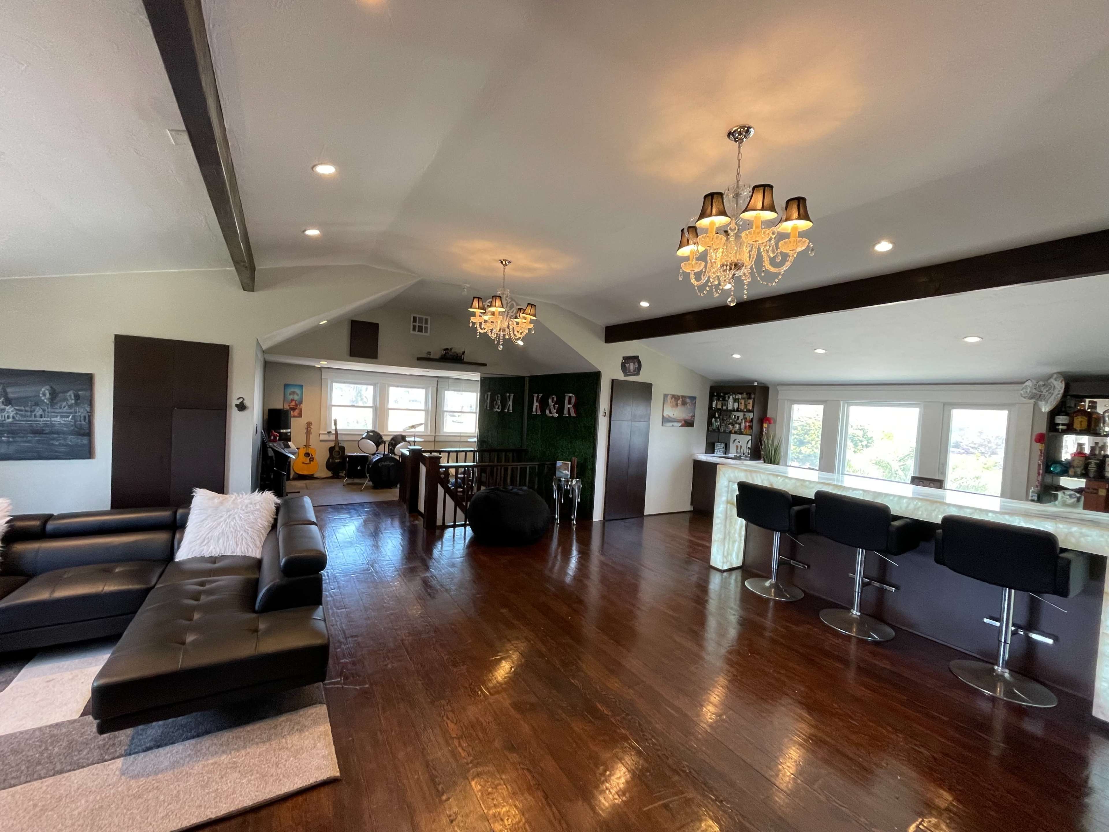 A modern living space featuring a black leather sectional sofa, a bar area with high stools, and decorative chandeliers hanging from the ceiling.