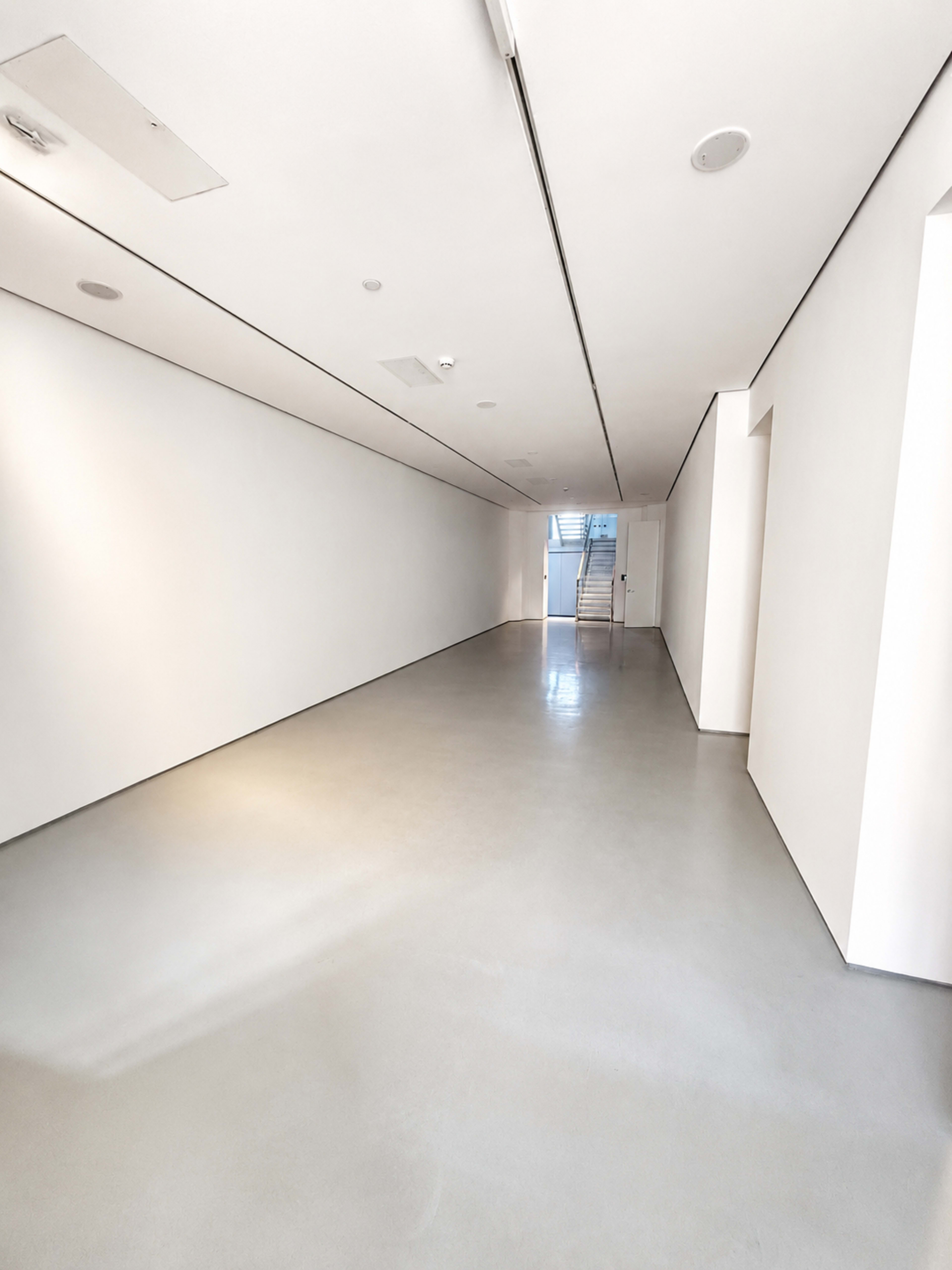 The image shows a long, narrow hallway with white walls and a smooth gray floor, leading to a set of stairs at the far end.