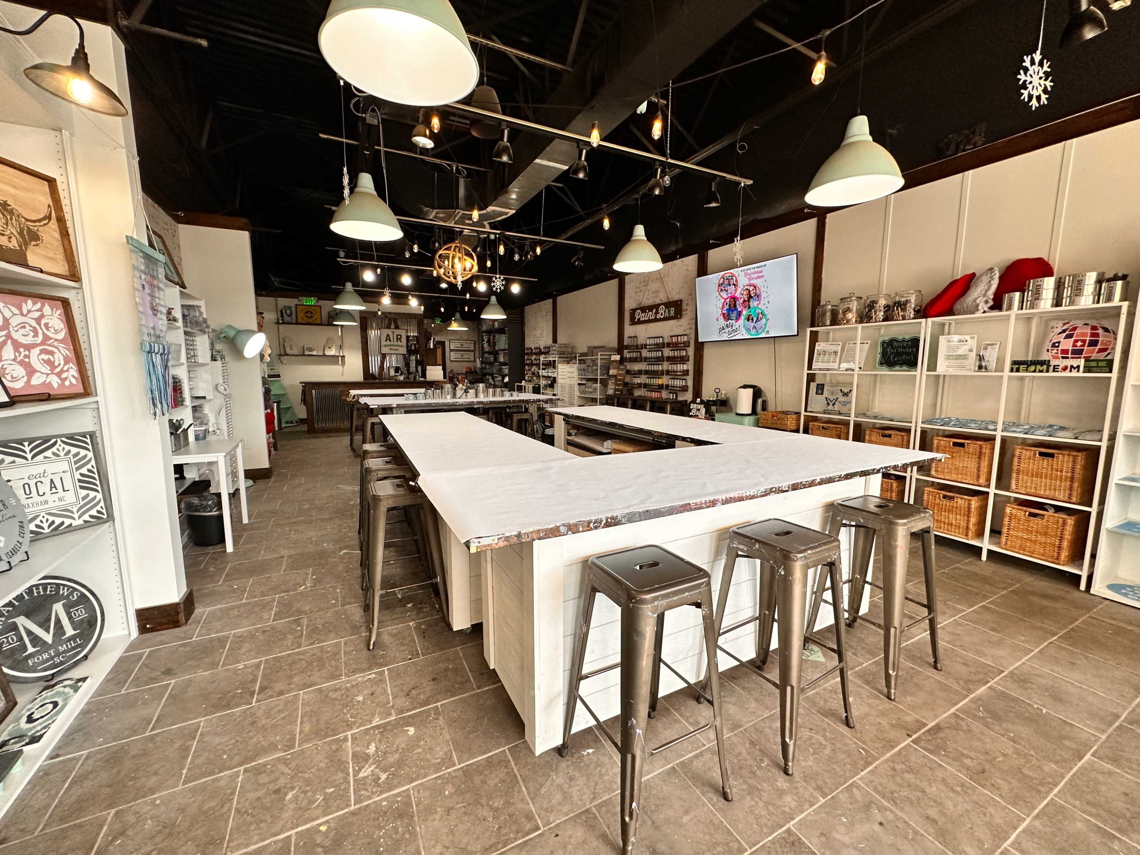 The image shows a spacious craft studio with several large tables, metal stools, and various shelves filled with craft supplies and decorations.