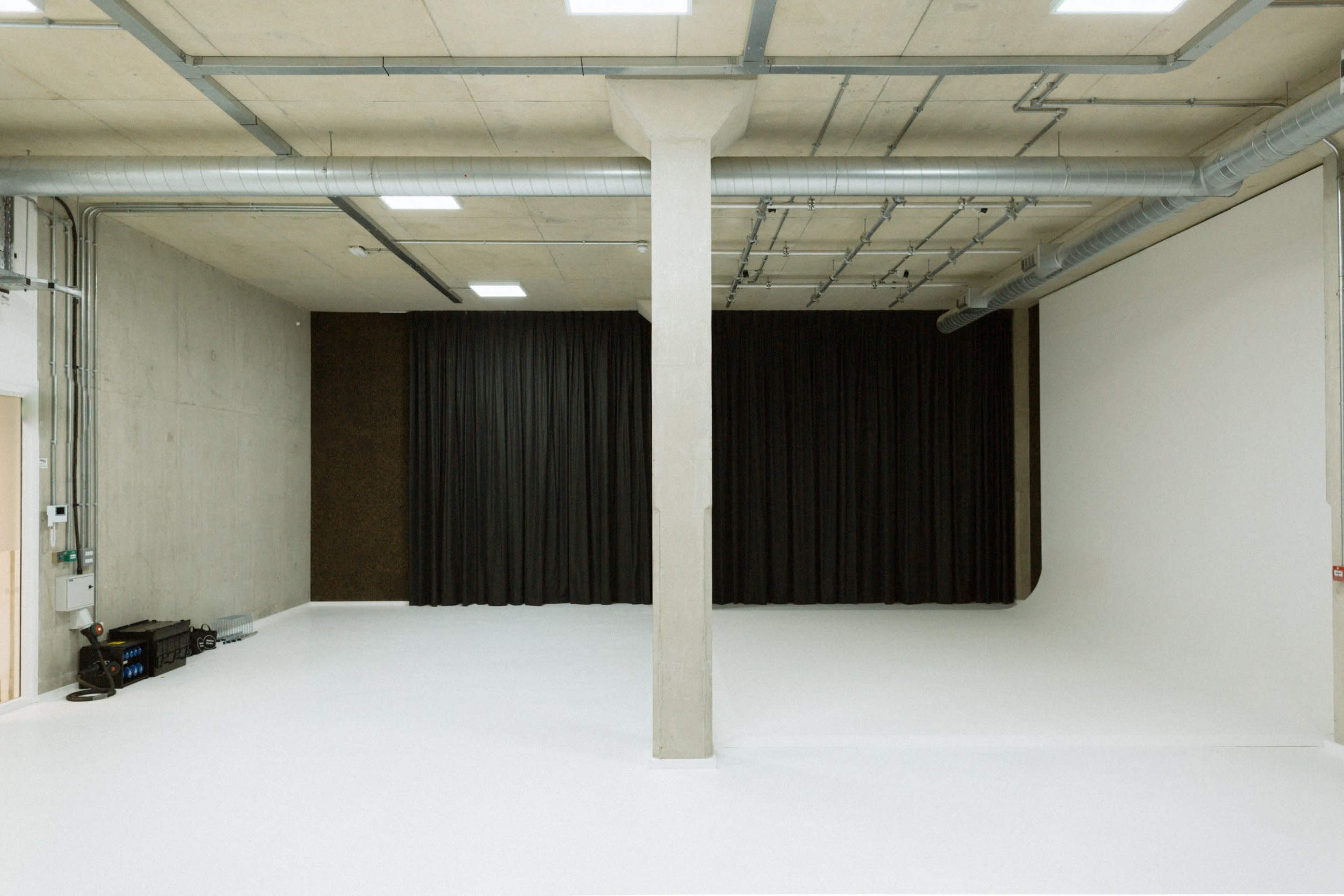 A spacious, minimalist studio with a white floor, a central concrete pillar, and black curtains on one side.