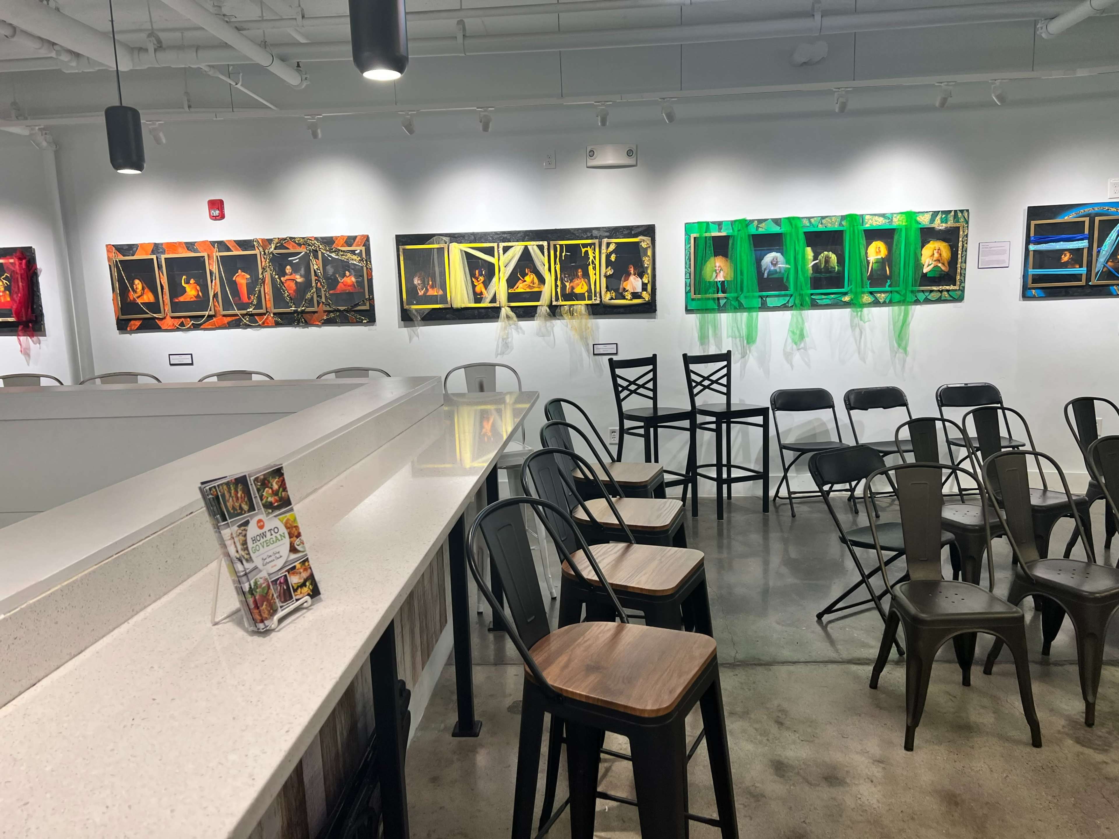 The image shows an art gallery with illuminated framed photographs hanging on the walls and empty chairs arranged in front of a countertop.