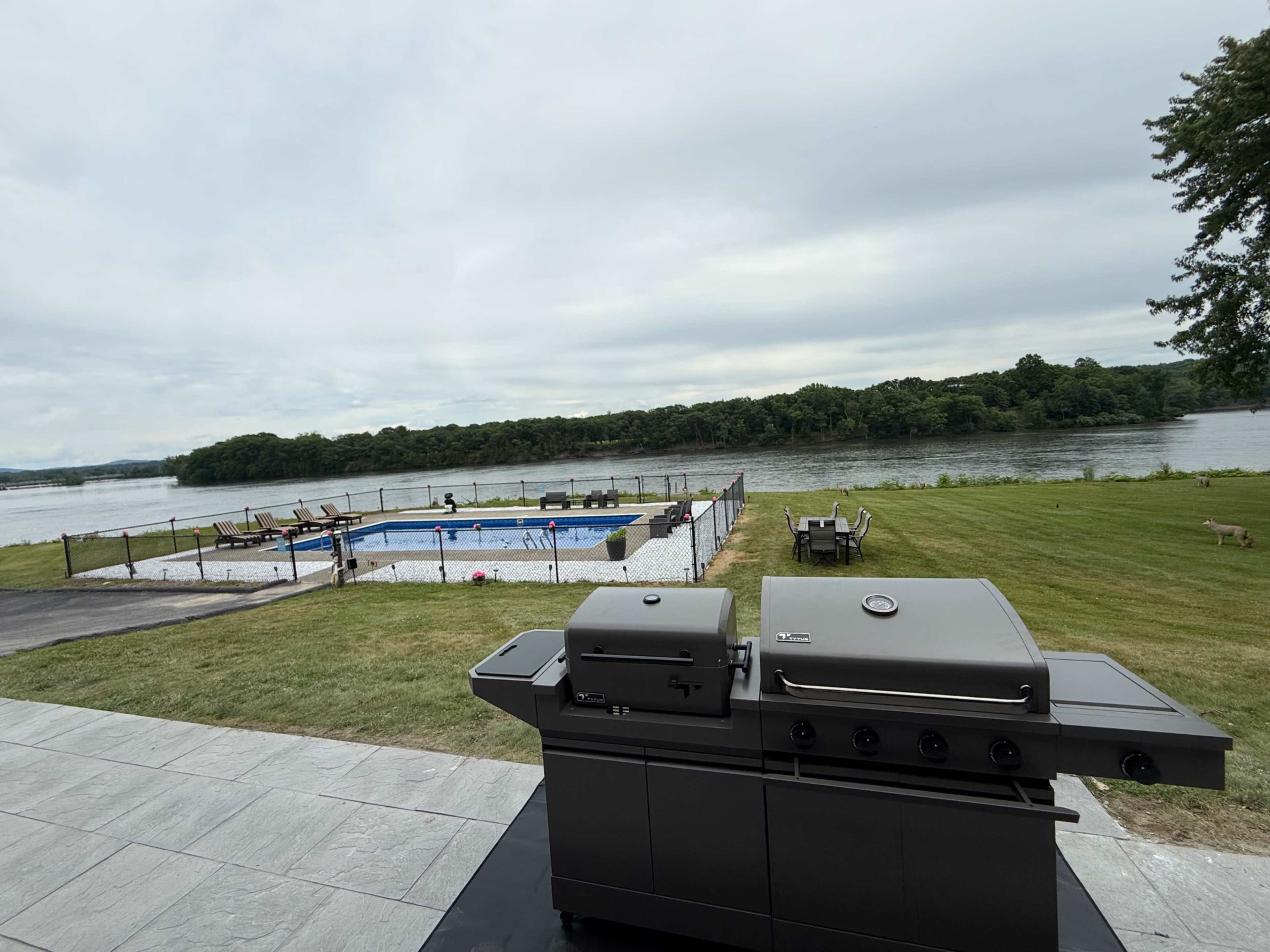 A stainless steel grill is positioned in the foreground, overlooking a pool area and a river bordered by trees in the background.