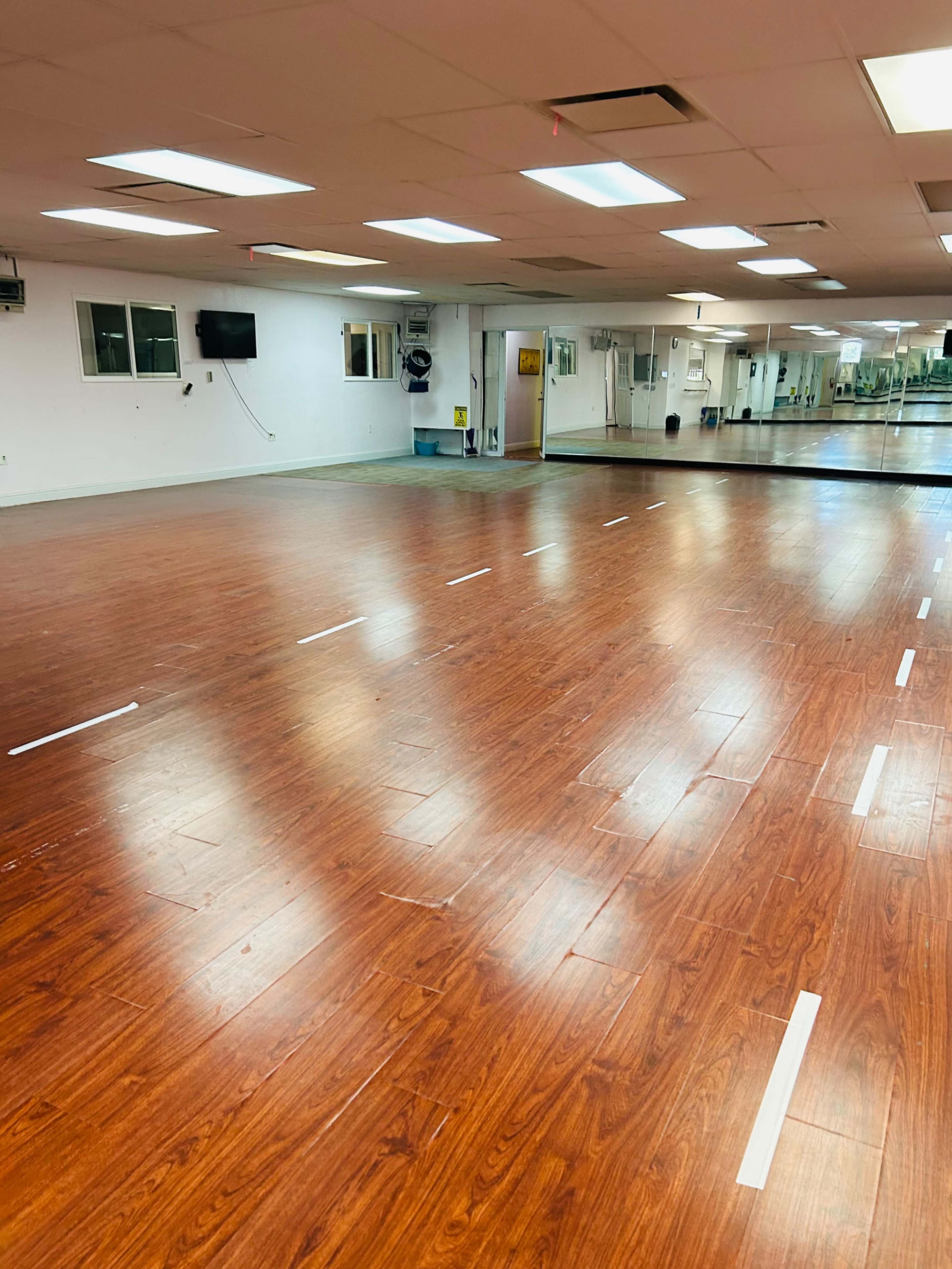 A spacious dance studio with a wooden floor and mirrored walls, featuring white markings on the floor.
