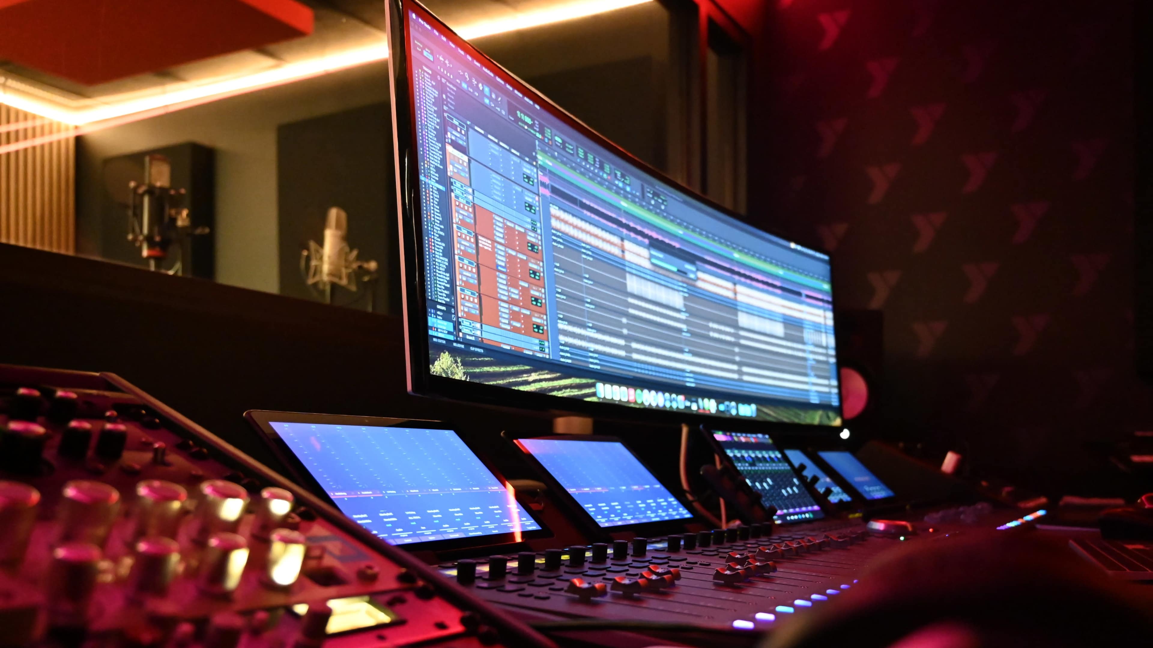 A music production studio with a large monitor displaying audio editing software and several control surfaces illuminated by soft lighting.