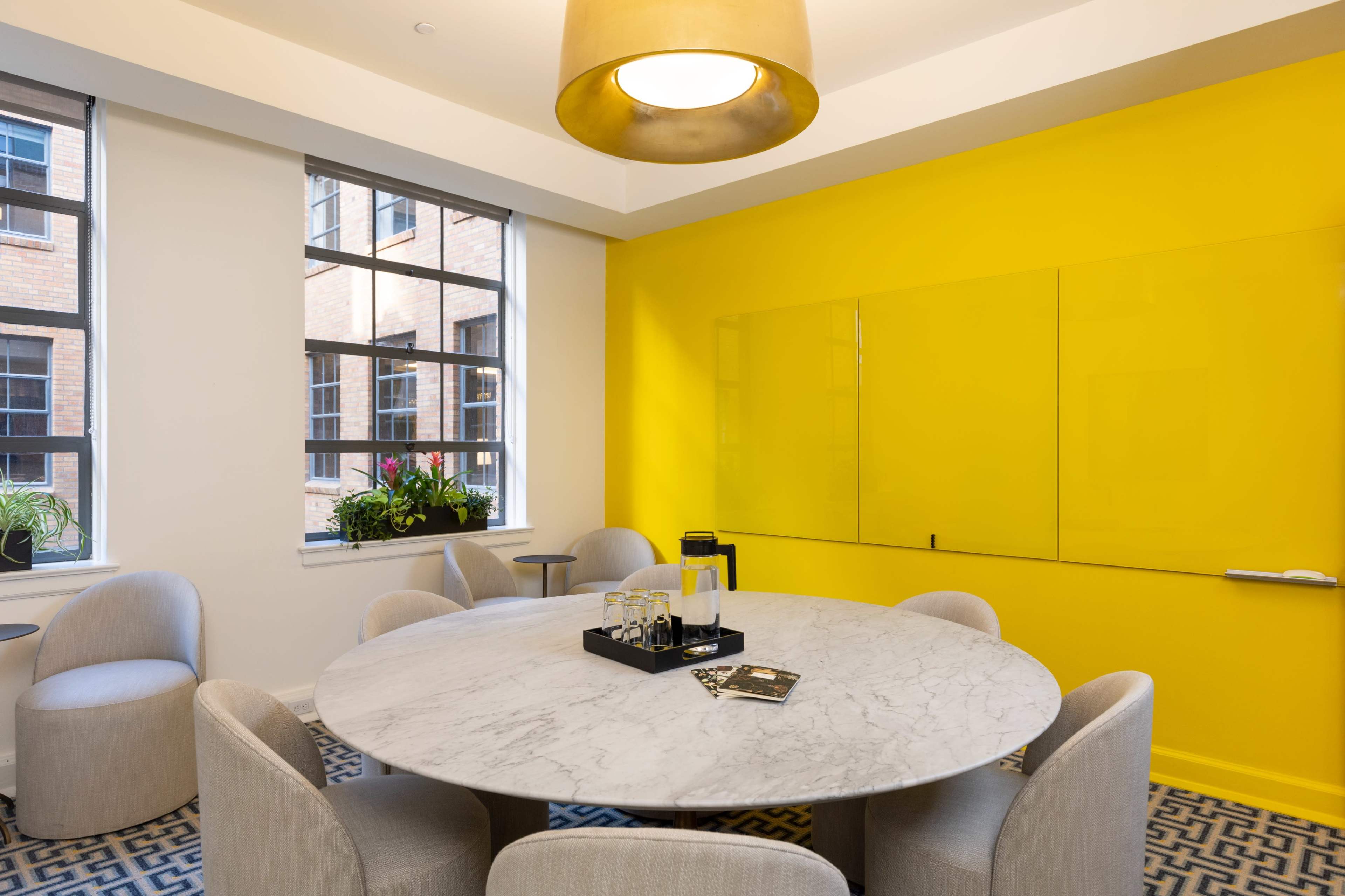 A round marble conference table with six grey chairs is set in a brightly painted yellow meeting room, featuring large windows and a glass whiteboard.