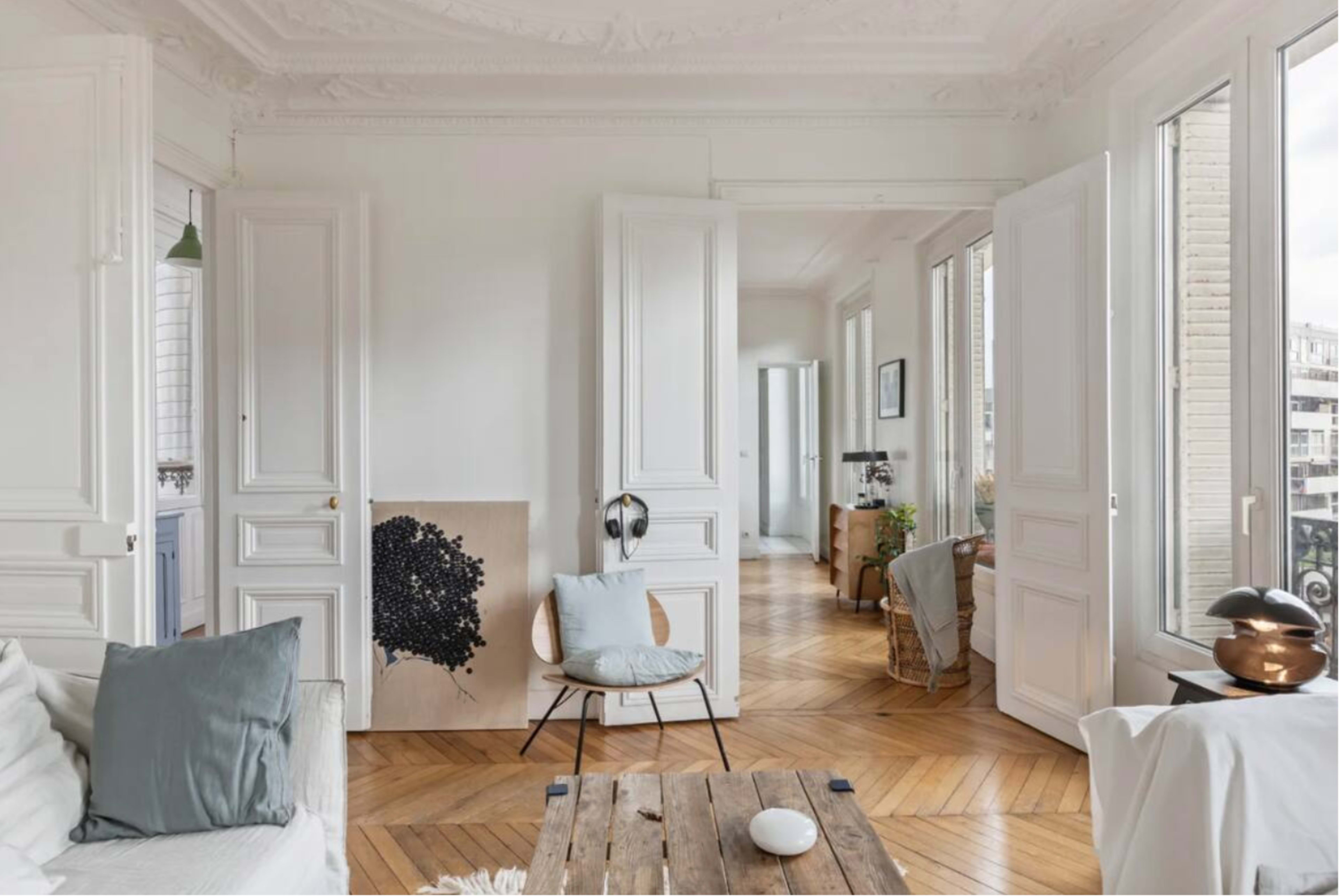 A bright, minimalistic living room features white walls, wooden herringbone flooring, a painting, and large windows allowing natural light.