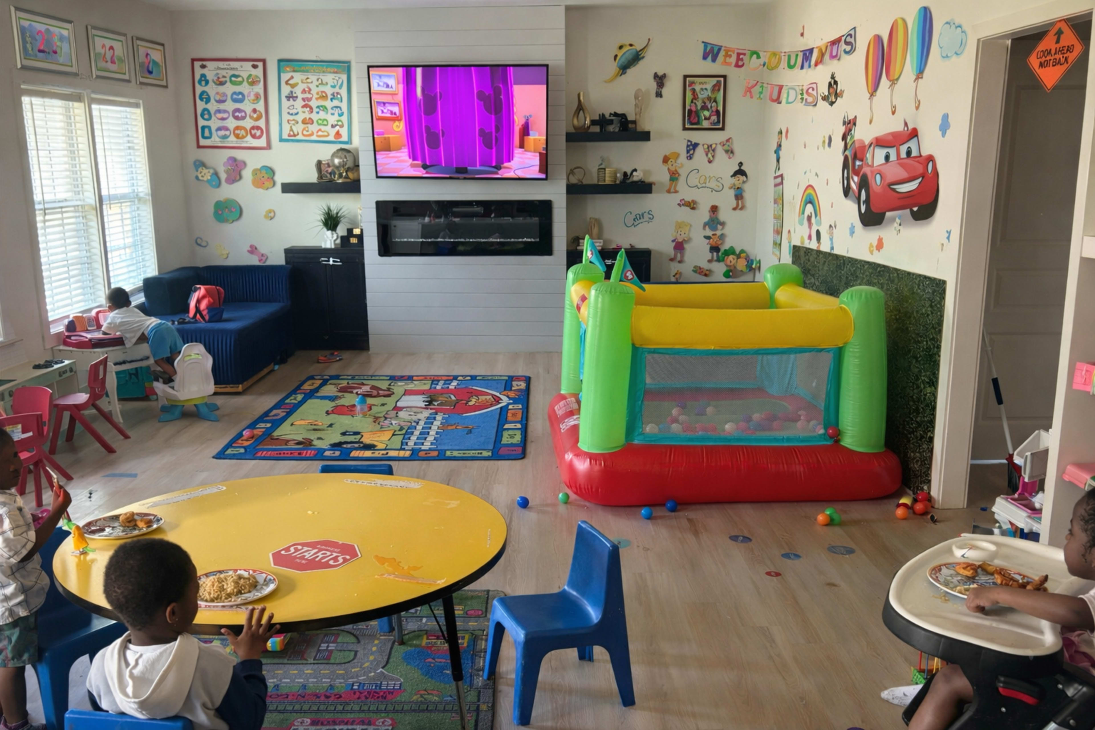 The image shows a vibrant children's playroom with colorful decor, a bouncy castle, a TV mounted on the wall, and children sitting at tables eating.