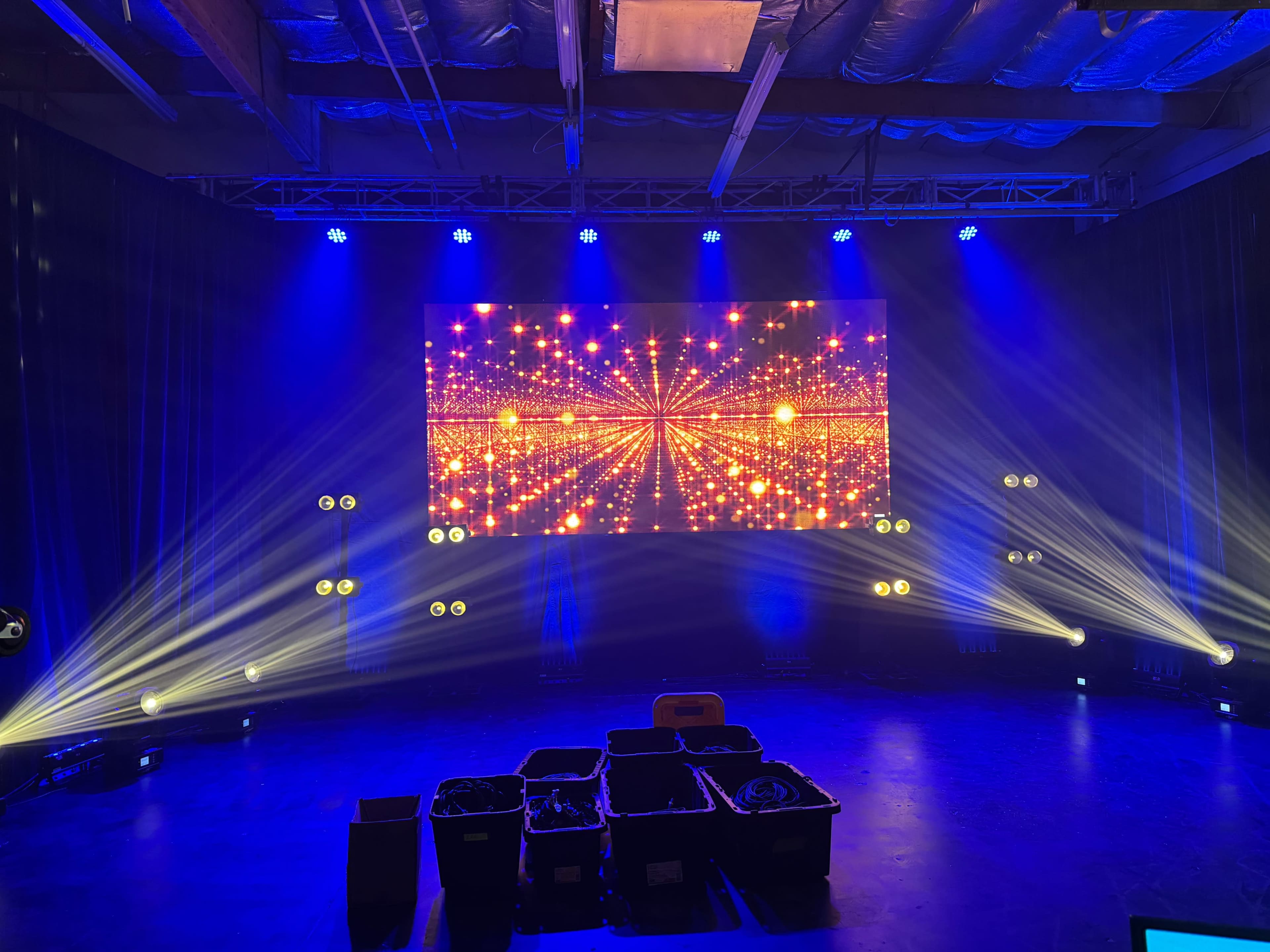 The scene features a stage setup with a large illuminated screen displaying vibrant graphics, surrounded by blue lighting and several spotlight fixtures.