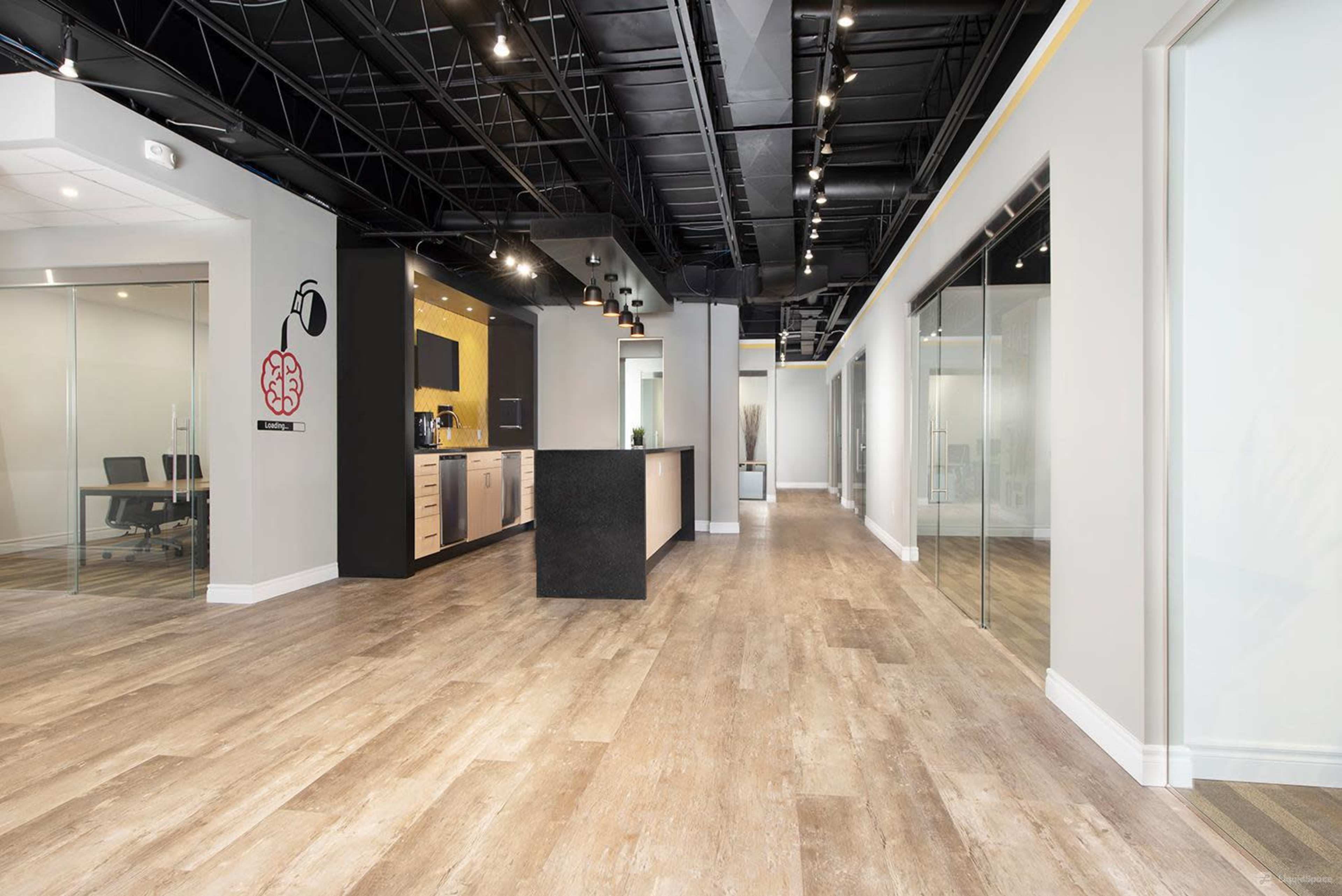 A modern office interior with a kitchen area on one side and glass-walled meeting rooms along the hallway.