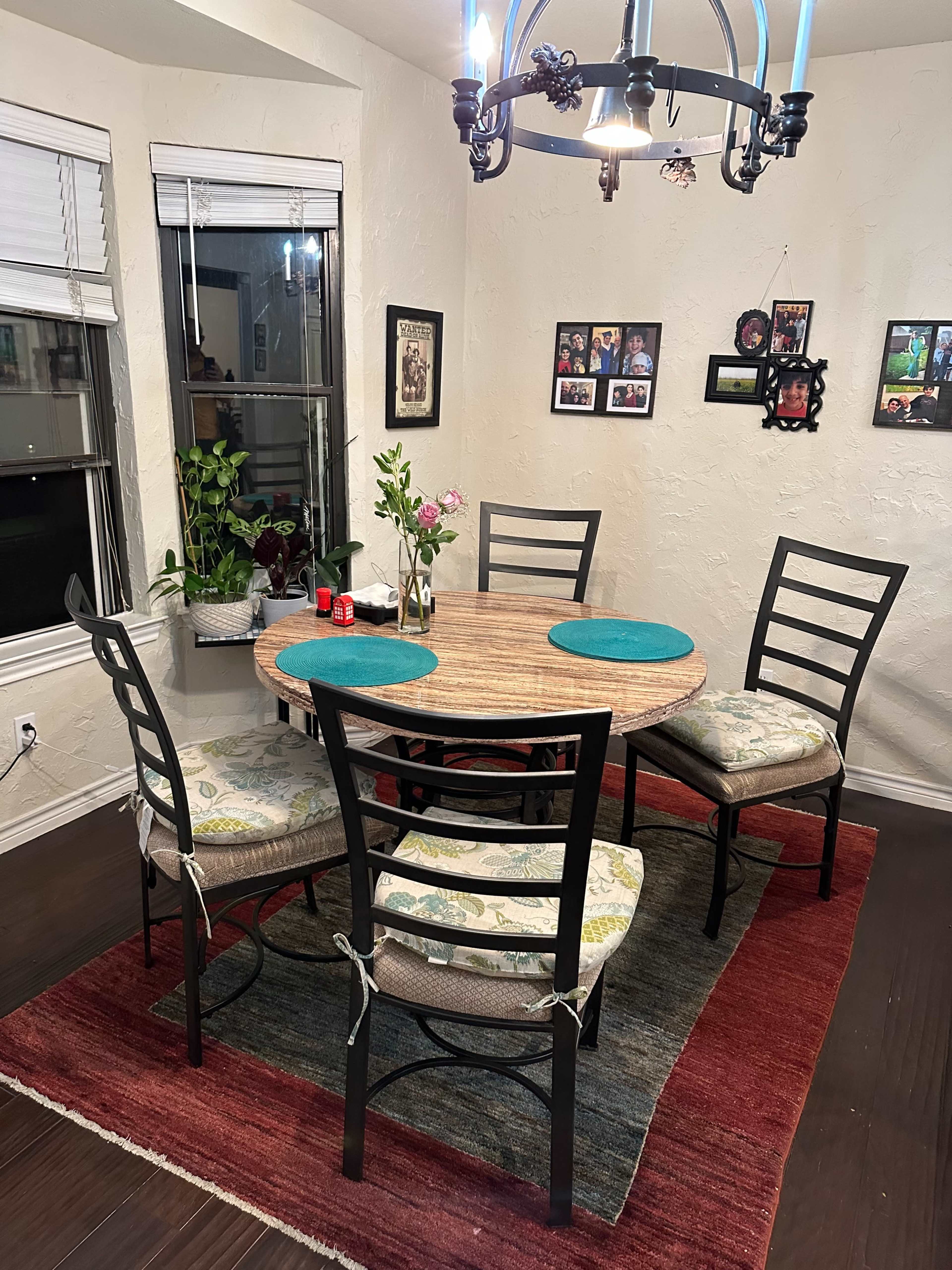 A round wooden dining table with four metal chairs, each featuring cushioned seats, is set in a room with a colorful rug and various plants, alongside framed photographs on the walls.