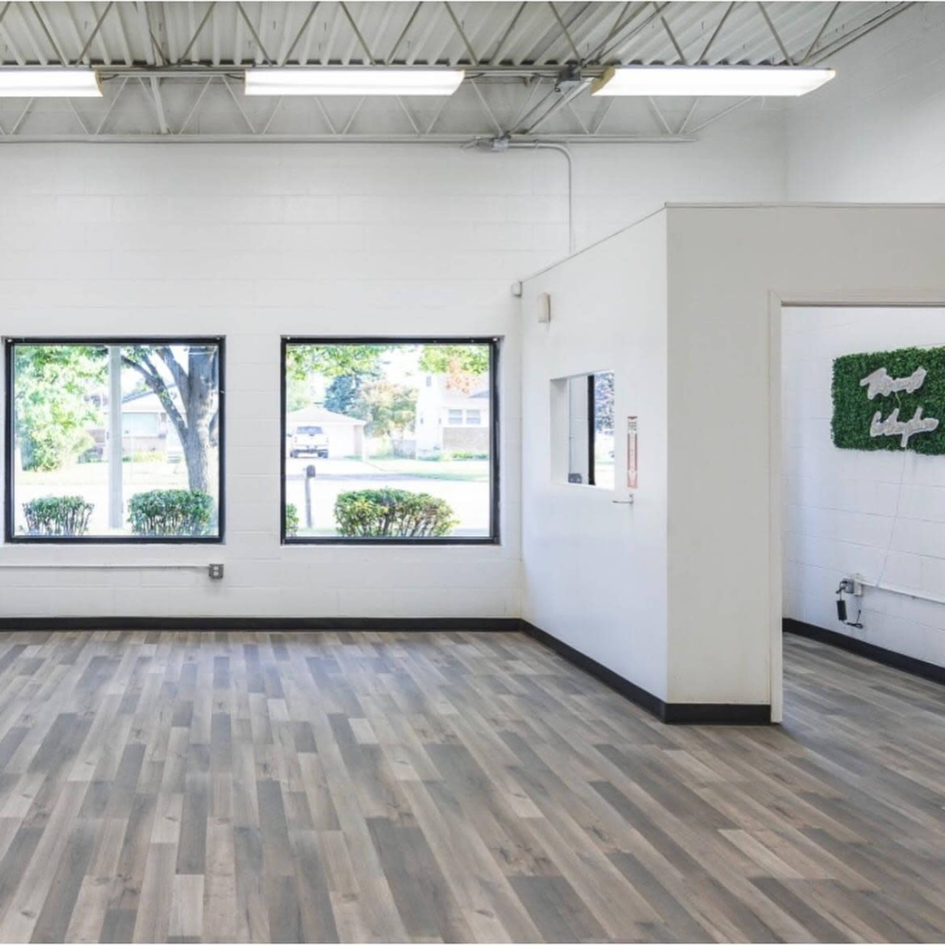 A spacious, empty room with large windows, light wood flooring, and a white wall with a doorway leading to another area.