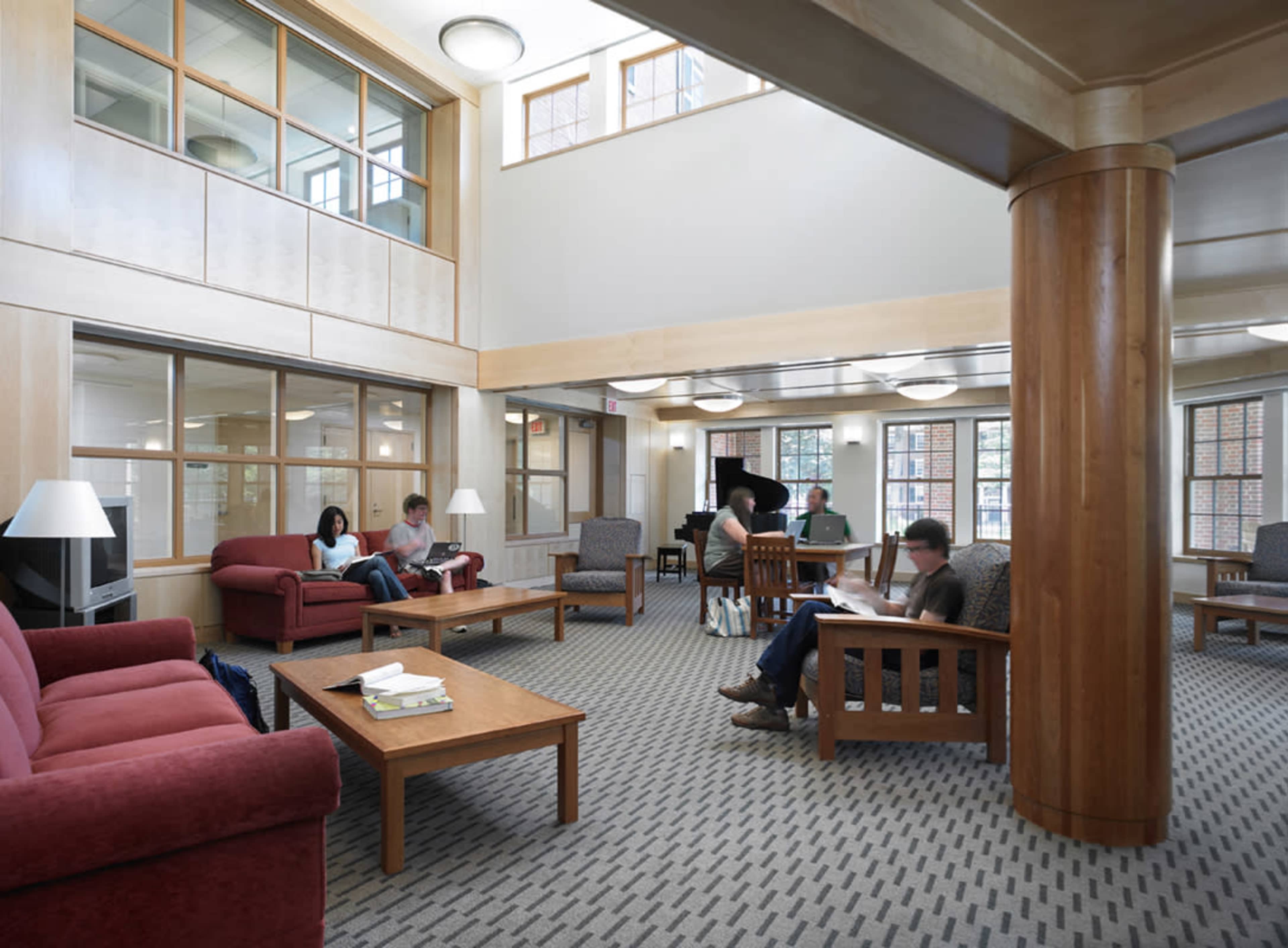 The image depicts a spacious, well-lit library lounge area with several people reading and studying, surrounded by comfortable seating and wooden furniture.