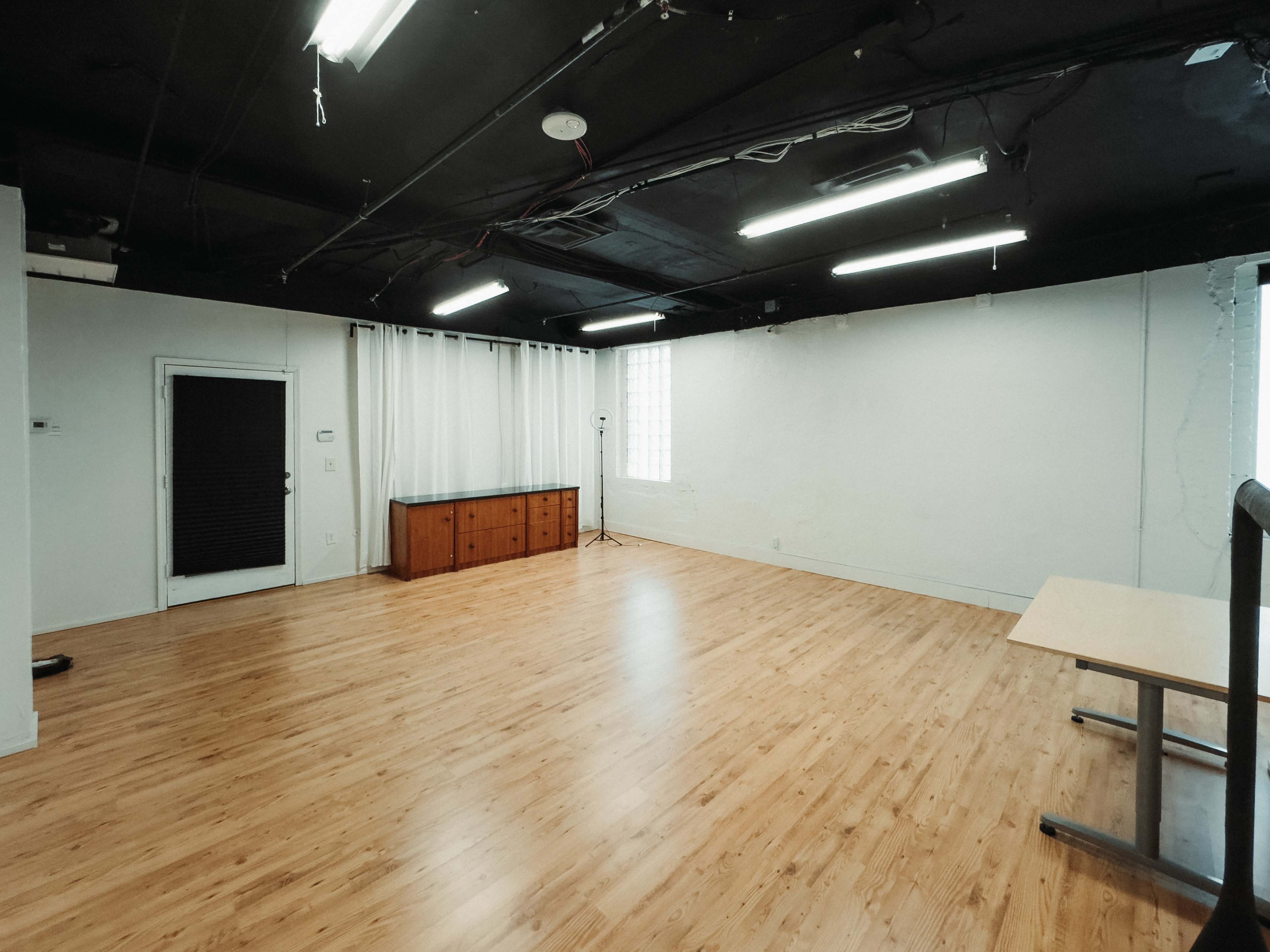 The image shows a spacious, empty room with wooden flooring, white walls, and two windows, illuminated by fluorescent lights.