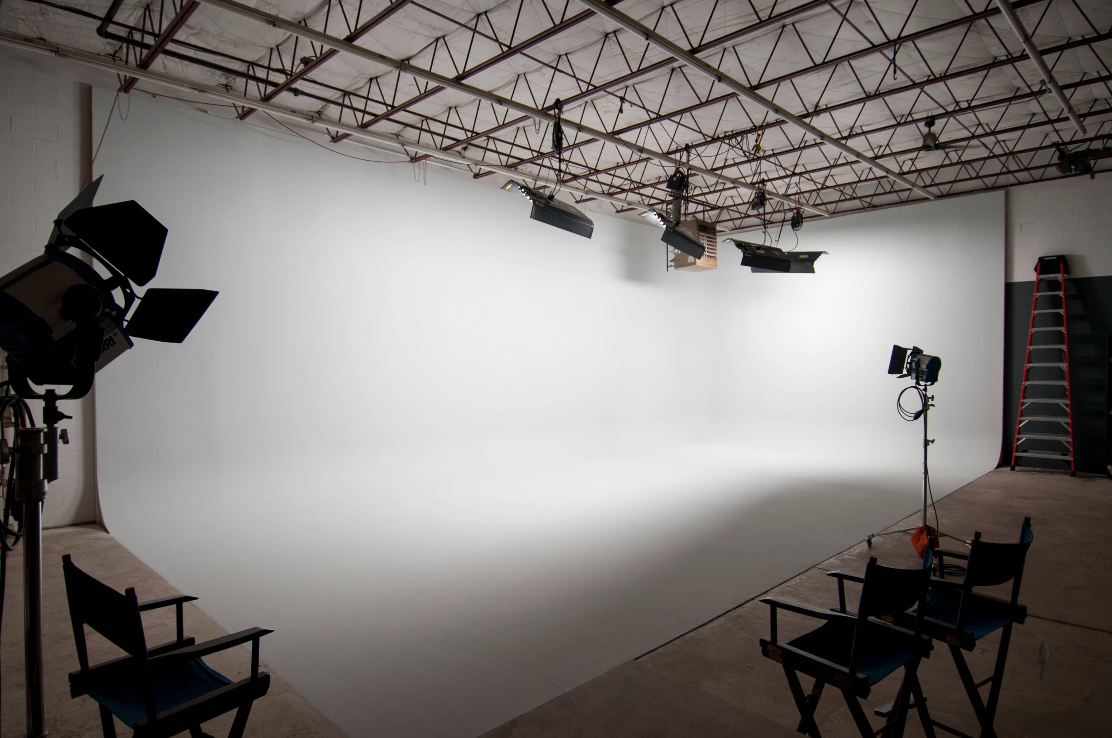 A photography studio features a seamless white backdrop, multiple lighting setups, and empty director's chairs.
