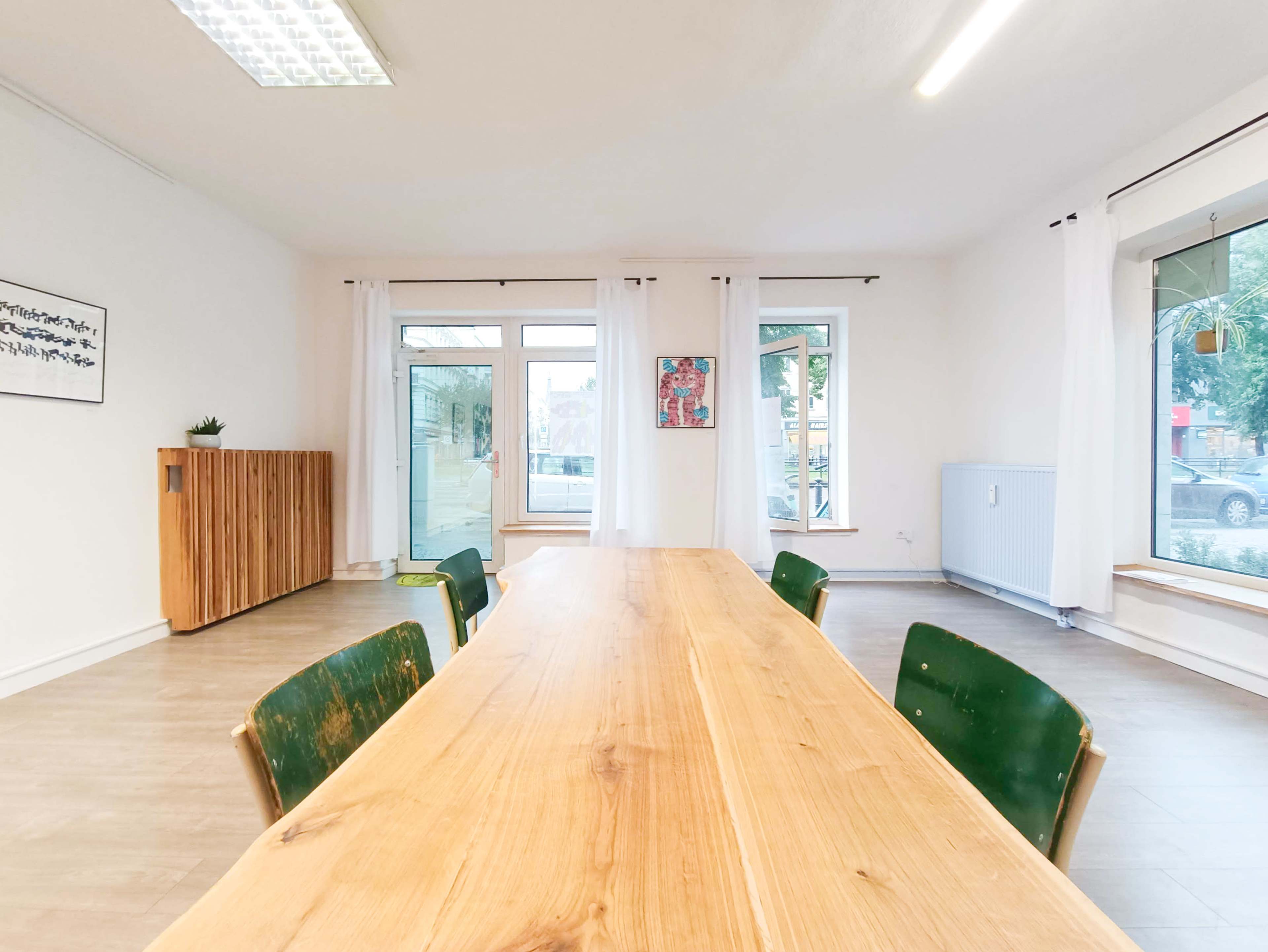 A long wooden table with green chairs is centered in a bright, minimalistic room featuring large windows and simple decor.